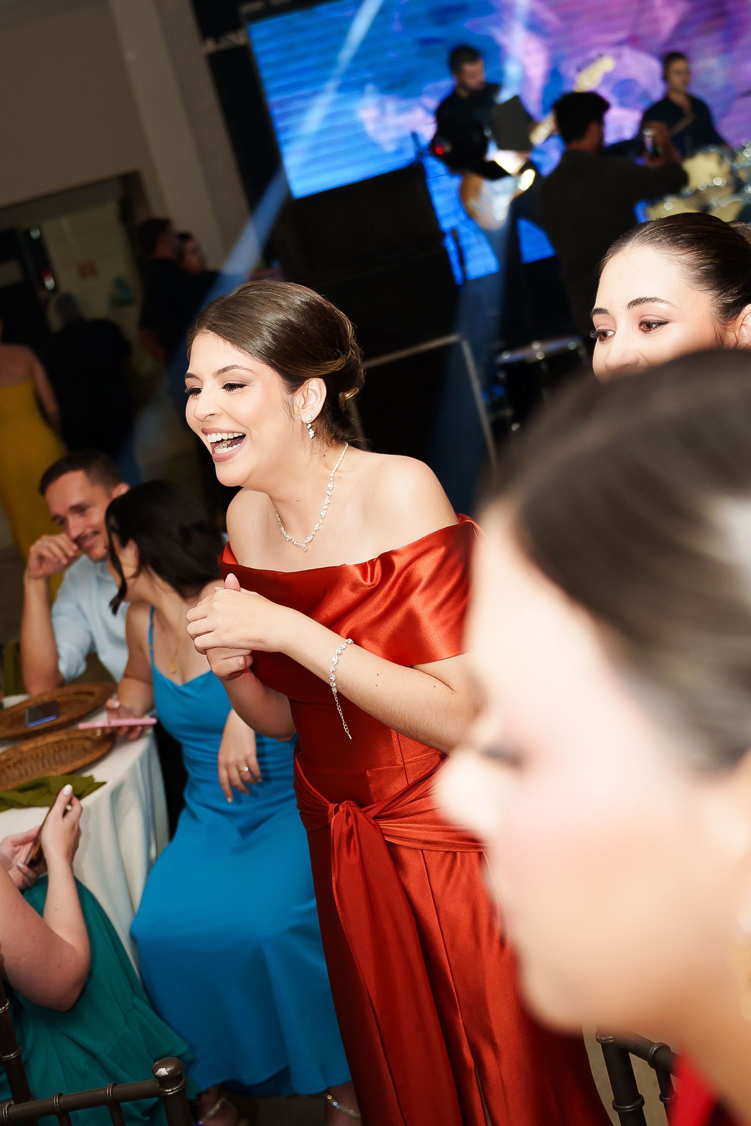 Women in elegant red and teal gowns laughing at a formal event with stage lighting in background.