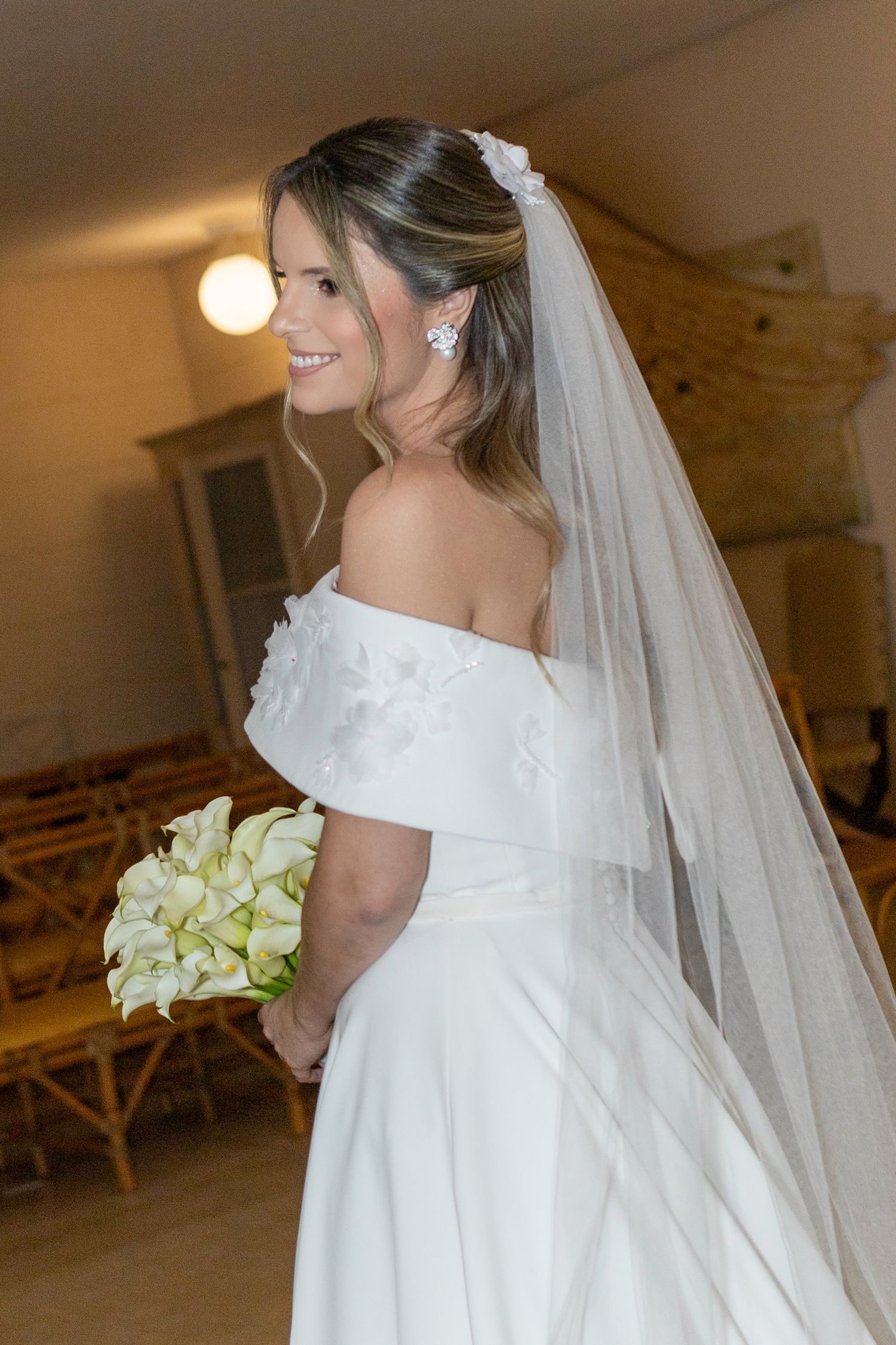 Smiling bride in off-shoulder white gown with veil holds calla lily bouquet inside a church venue.