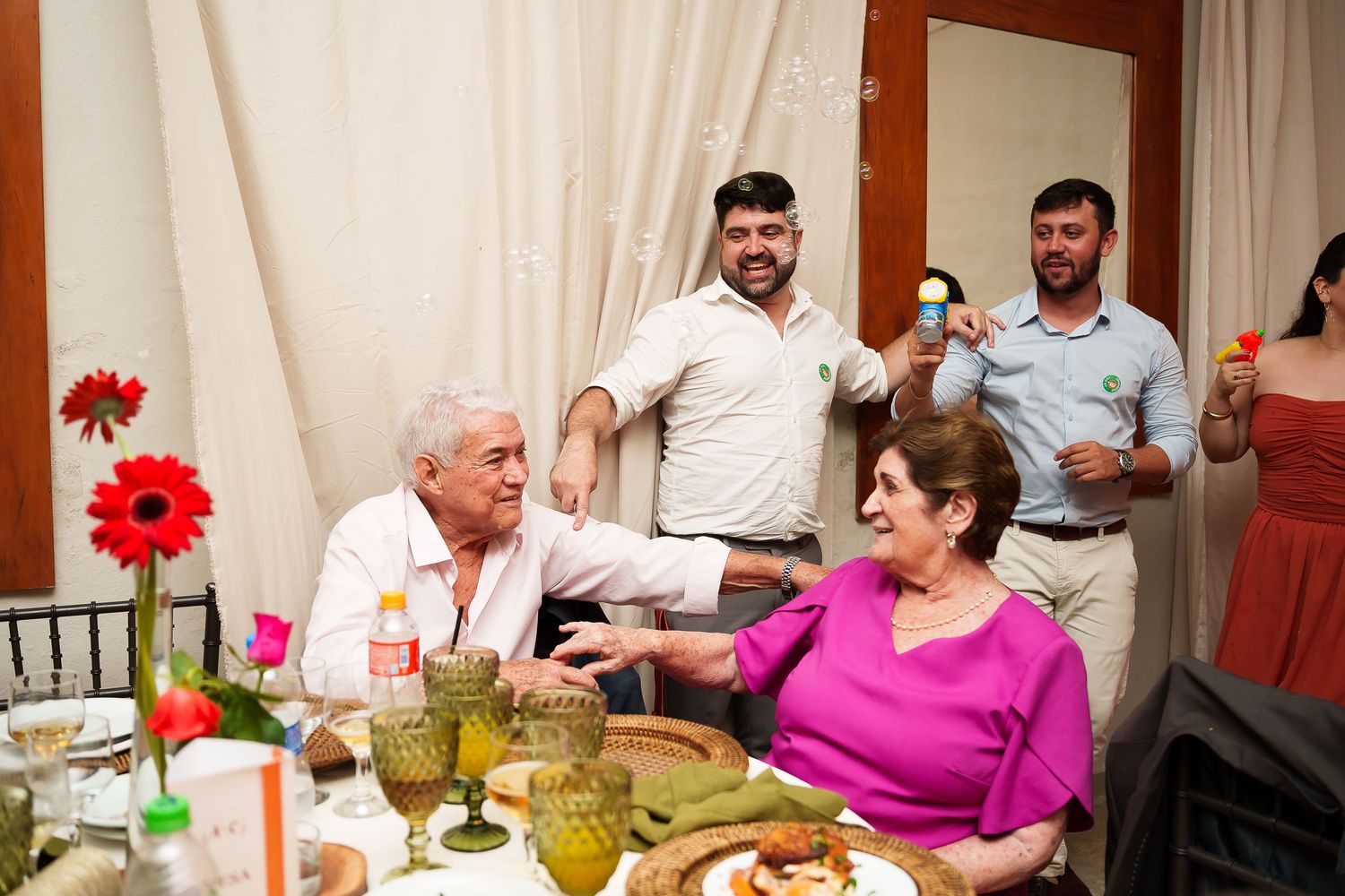 Family gathering around a festive dinner table with red flowers, raising glasses in a celebratory toast indoors.