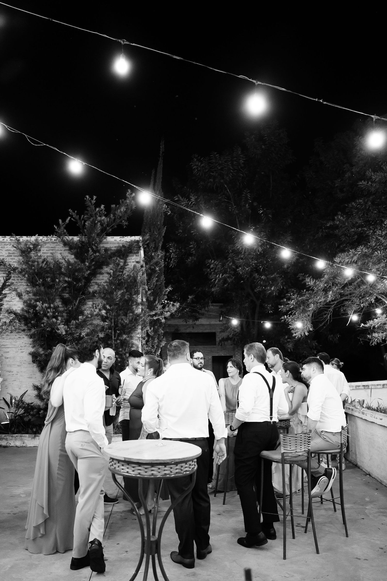 Black and white photo of wedding guests mingling outdoors at night under string lights at an elegant evening reception.