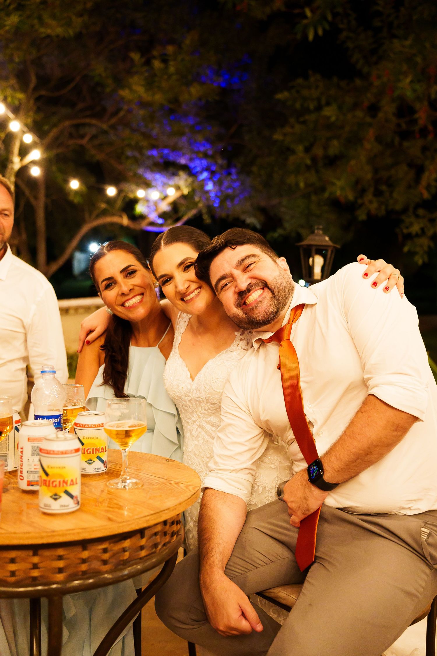 Three friends smile and pose together at an outdoor evening wedding reception with string lights.