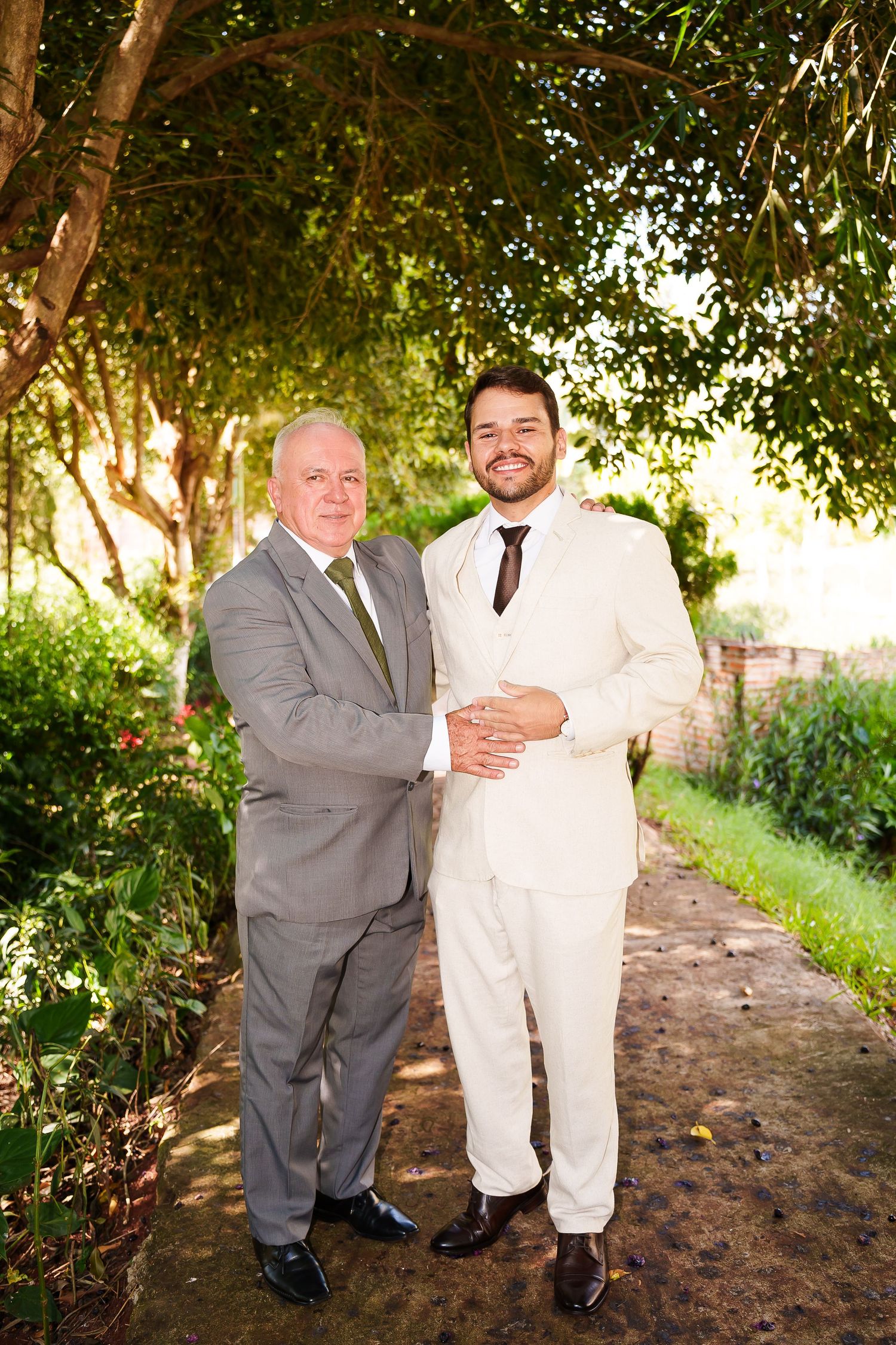 Two formally dressed men in suits pose together outdoors on a garden path surrounded by lush green trees.