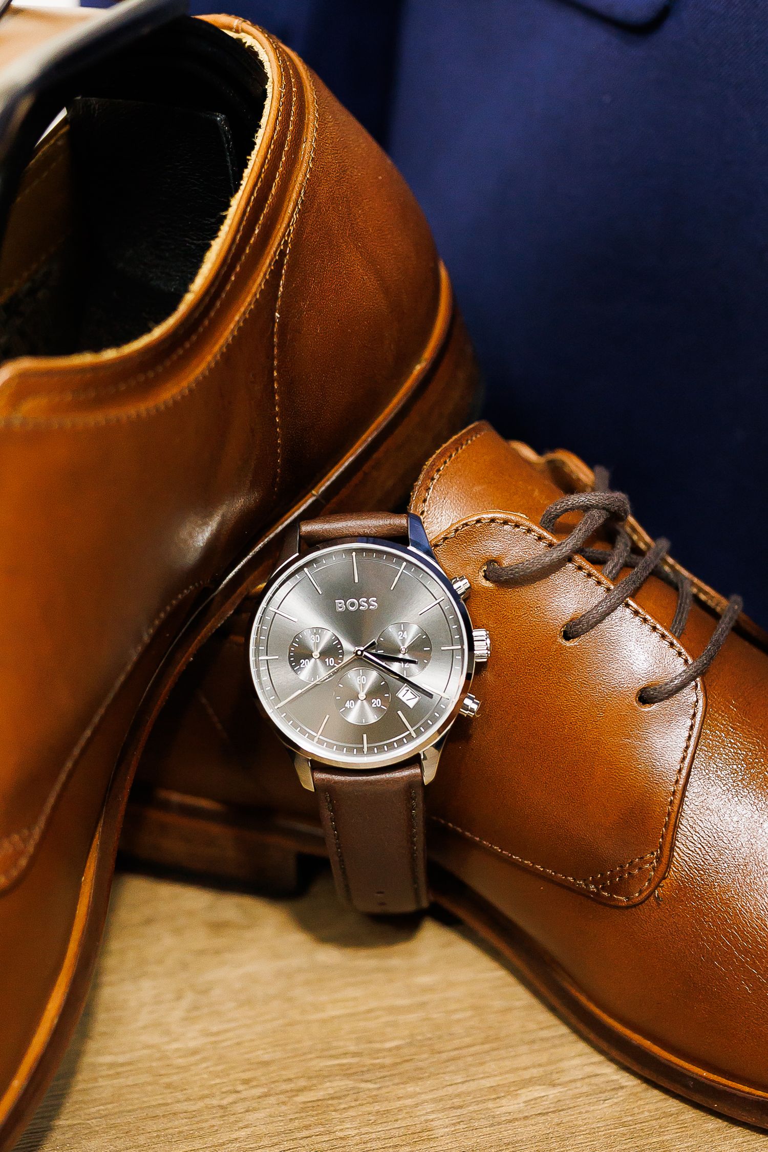Hugo Boss chronograph watch with brown leather strap displayed against tan leather Oxford dress shoes on wooden surface.