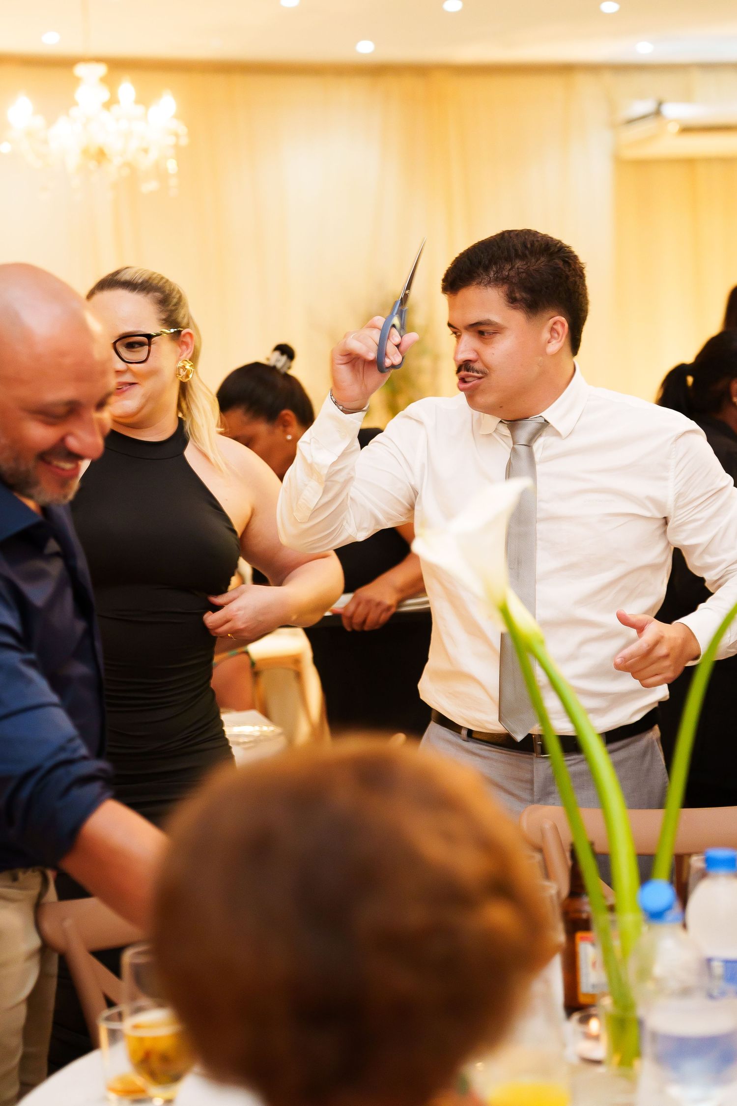 Man in white shirt and tie raises phone at elegant banquet hall gathering with guests in formal attire.