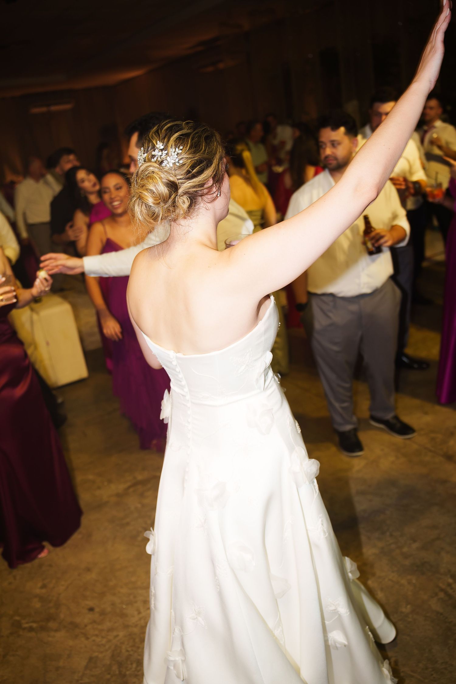 Bride in white backless gown dancing with raised arm at wedding reception, guests in purple bridesmaid dresses.