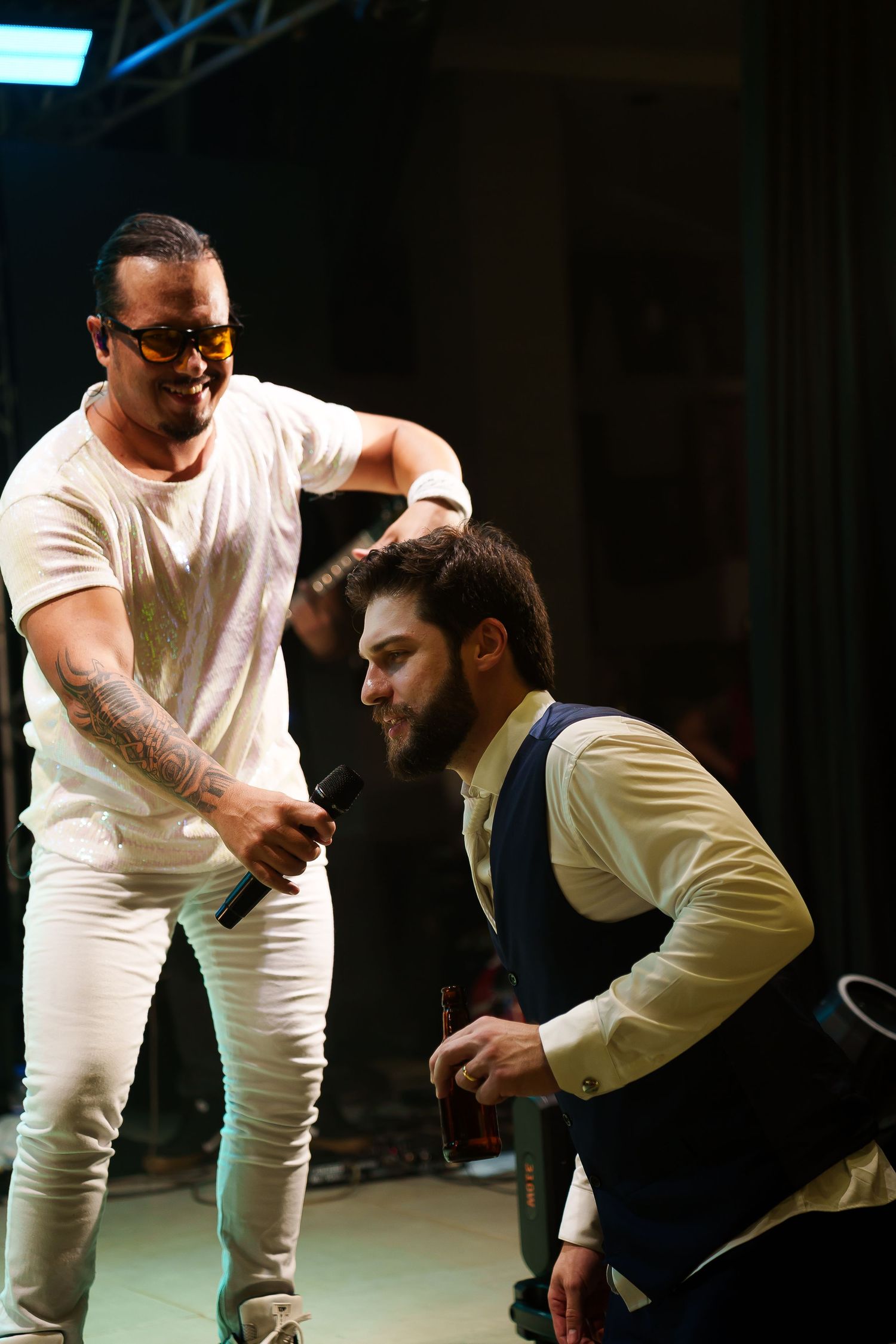 Two men on stage, one holding a microphone in white outfit, the other kneeling with a bottle, at a live event.