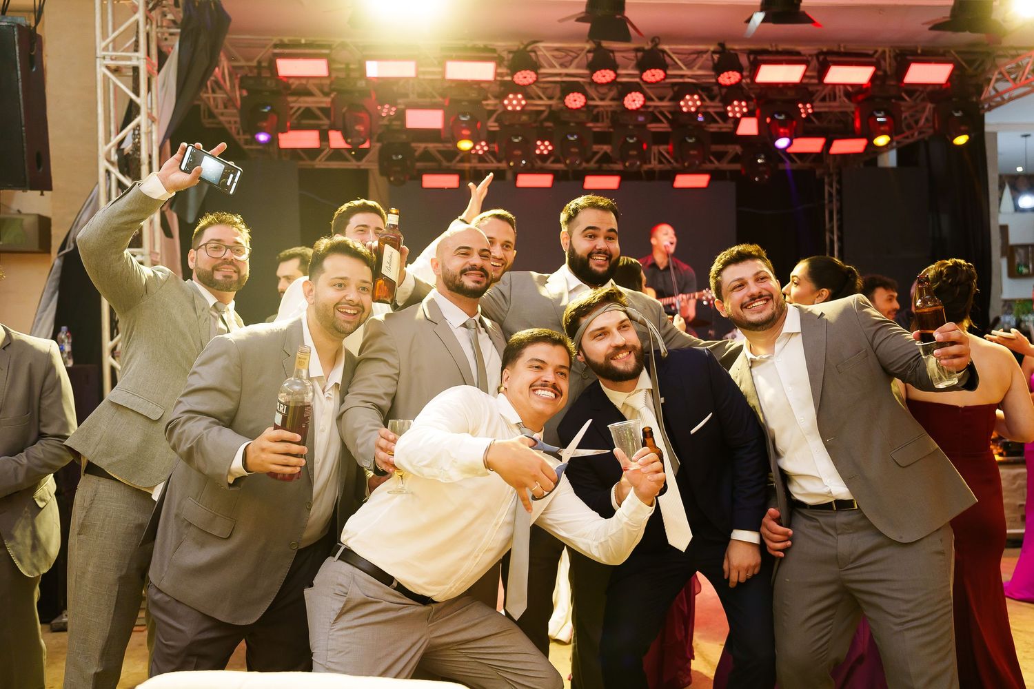 Groom and groomsmen taking a fun selfie together at a wedding reception venue with stage lighting.