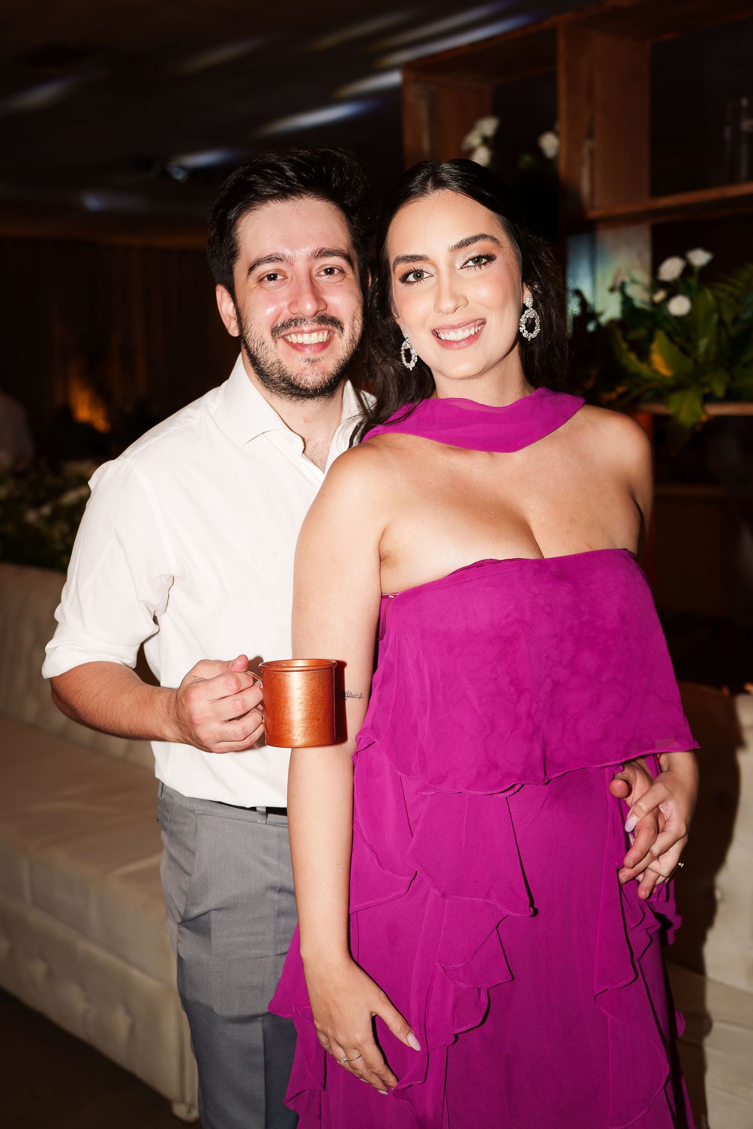 A smiling couple poses at a formal event, man in white shirt holding copper mug, woman in elegant magenta strapless gown.