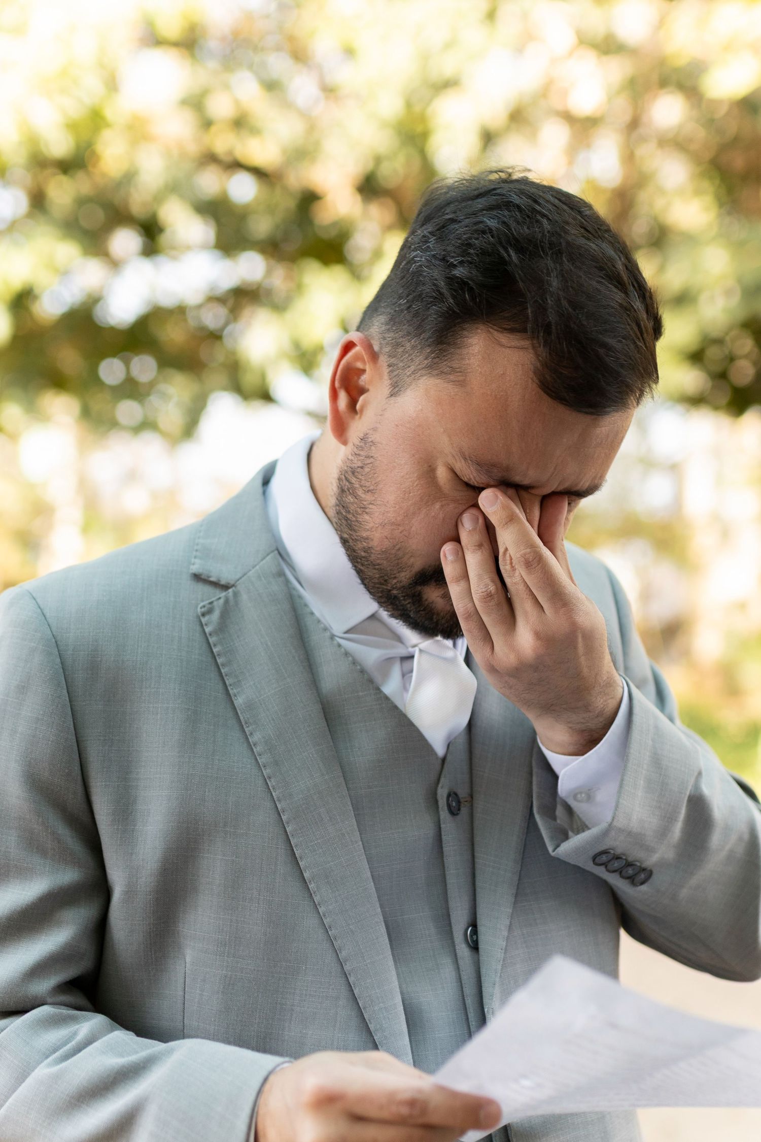 Upset bearded businessman in gray suit covering his eyes while holding a document outdoors.