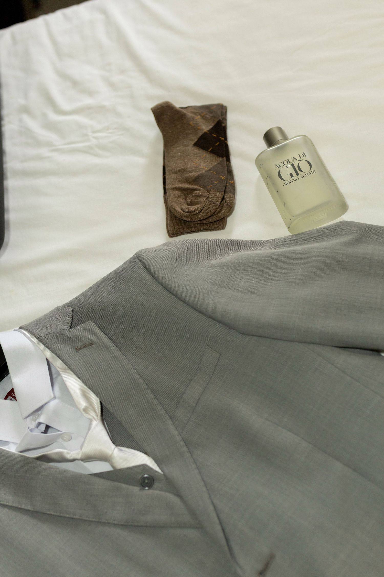 Gray suit jacket with white dress shirt, brown patterned socks, and Acqua Di Gio cologne on white bed.