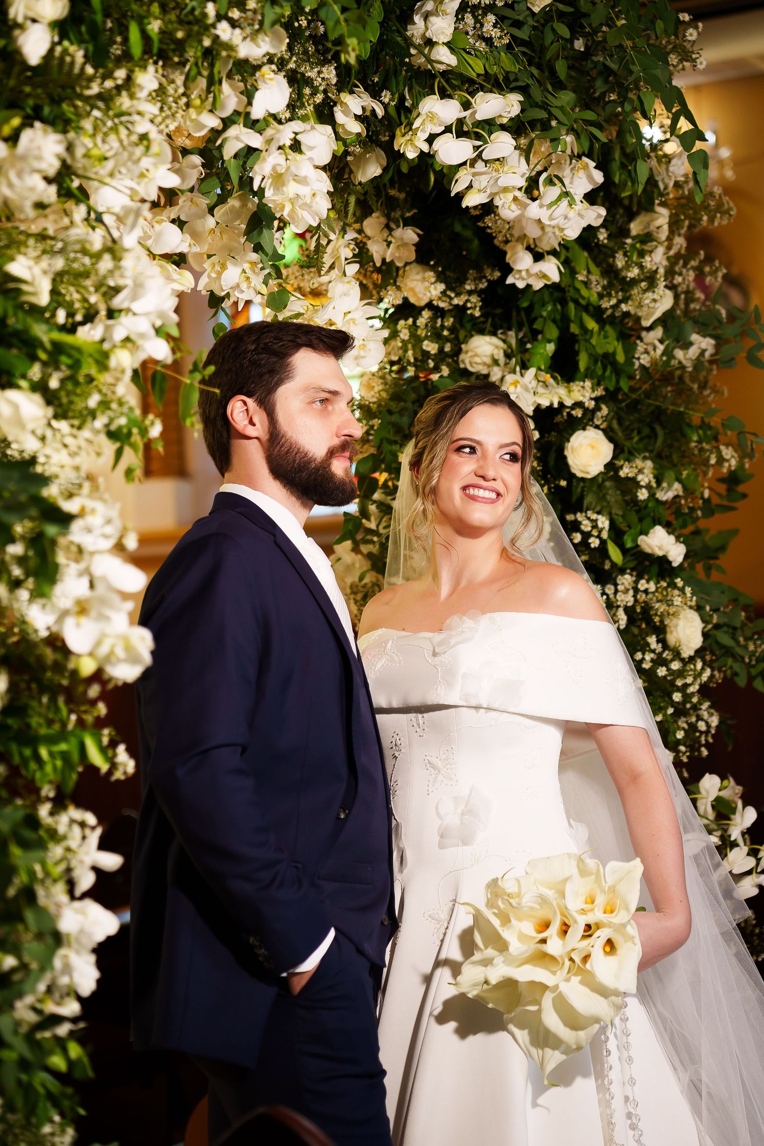 Bride in white off-shoulder gown holding calla lily bouquet stands with bearded groom in navy suit under floral arch.