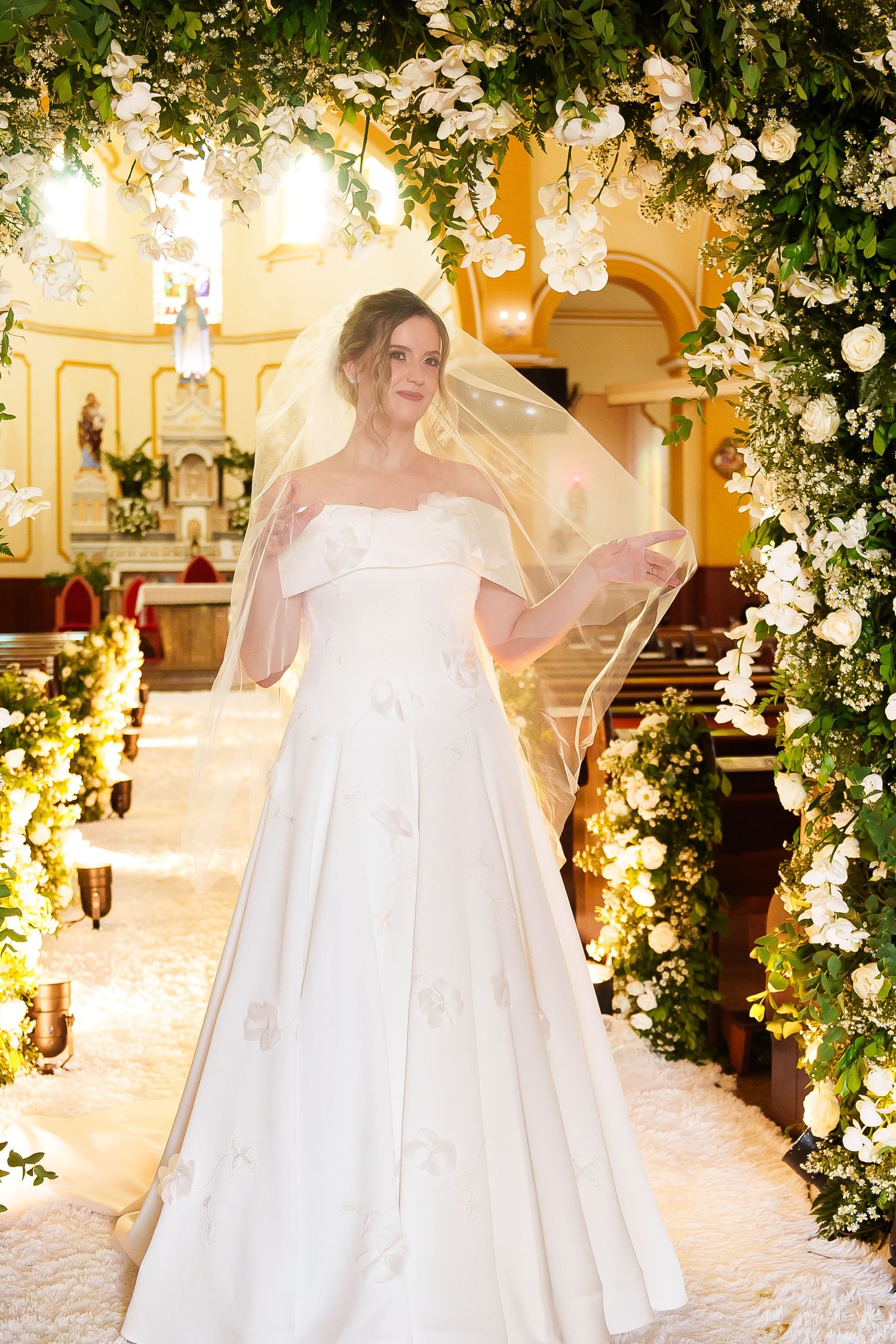 Bride in off-shoulder white ball gown with veil stands under floral arch in elegantly decorated church.