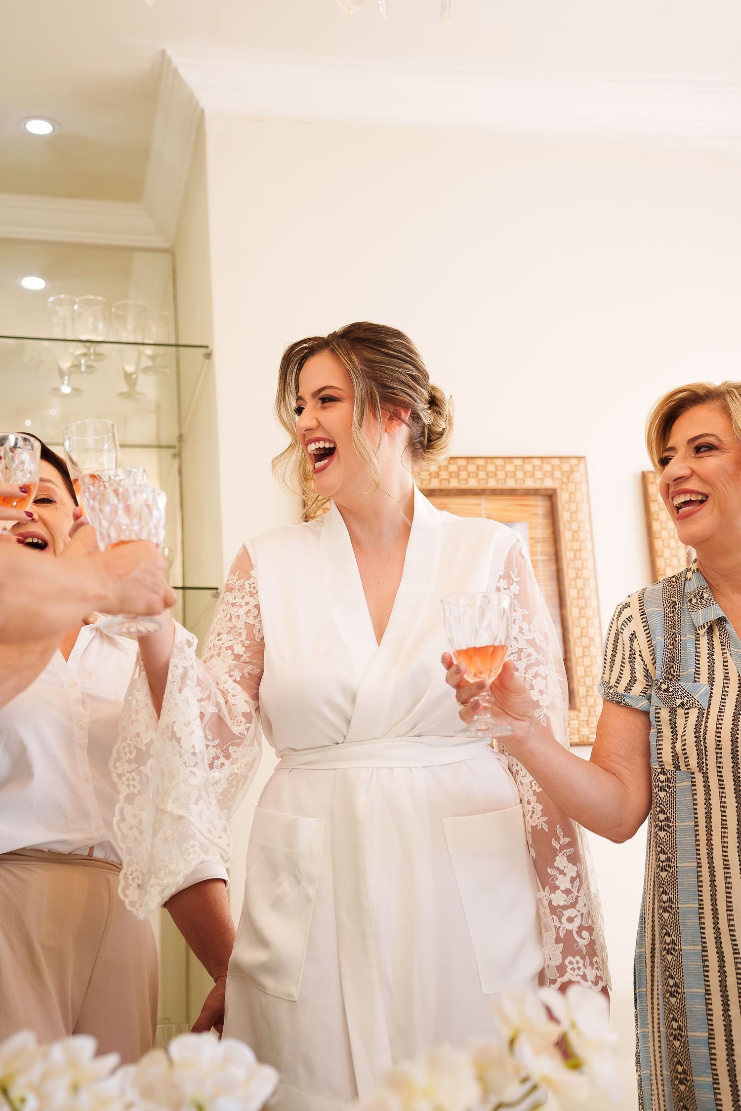 Bride in white lace robe laughing with bridesmaids holding champagne glasses during wedding preparations.