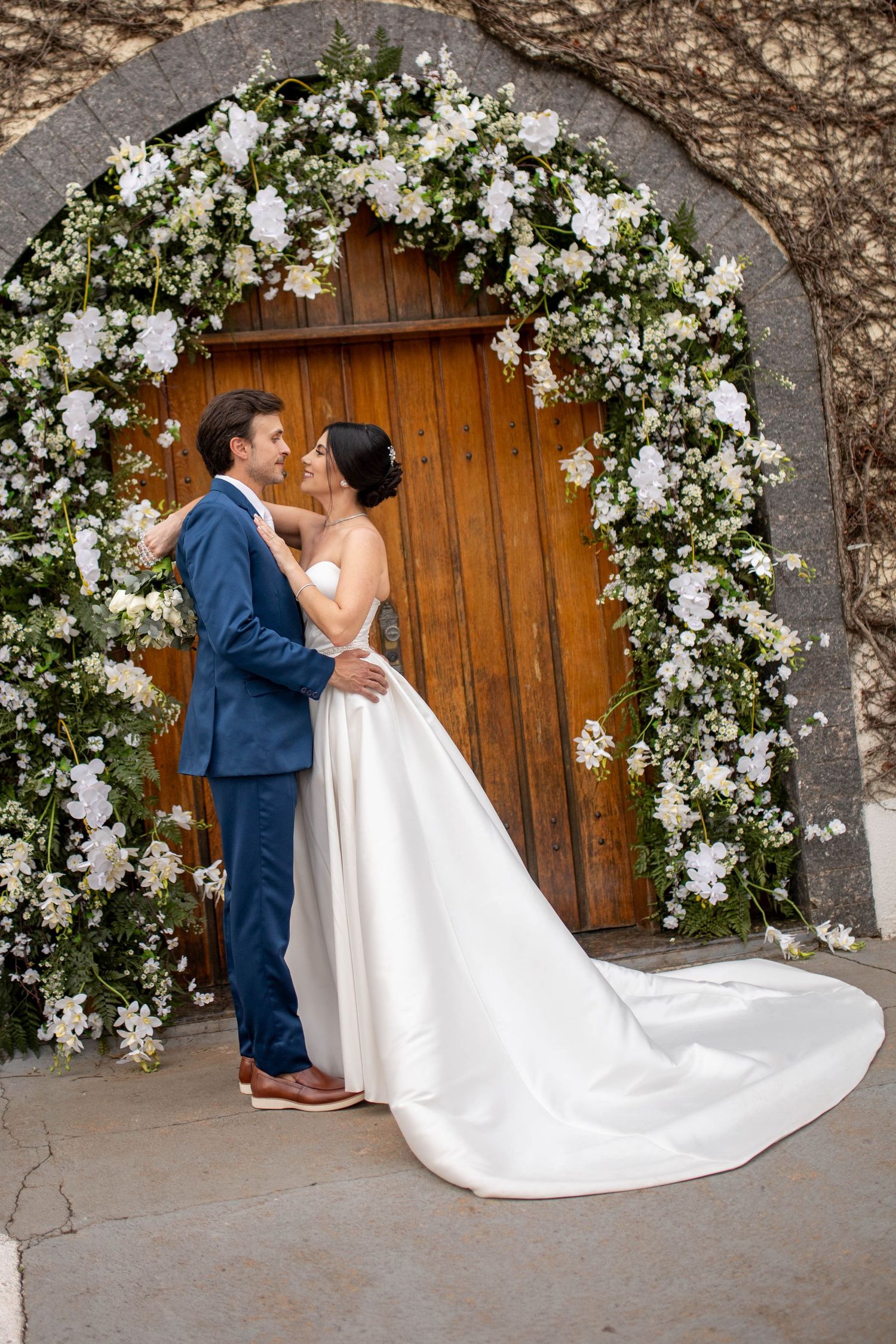 Bride and groom embrace under a floral arch of white flowers at a wooden door on their wedding day.