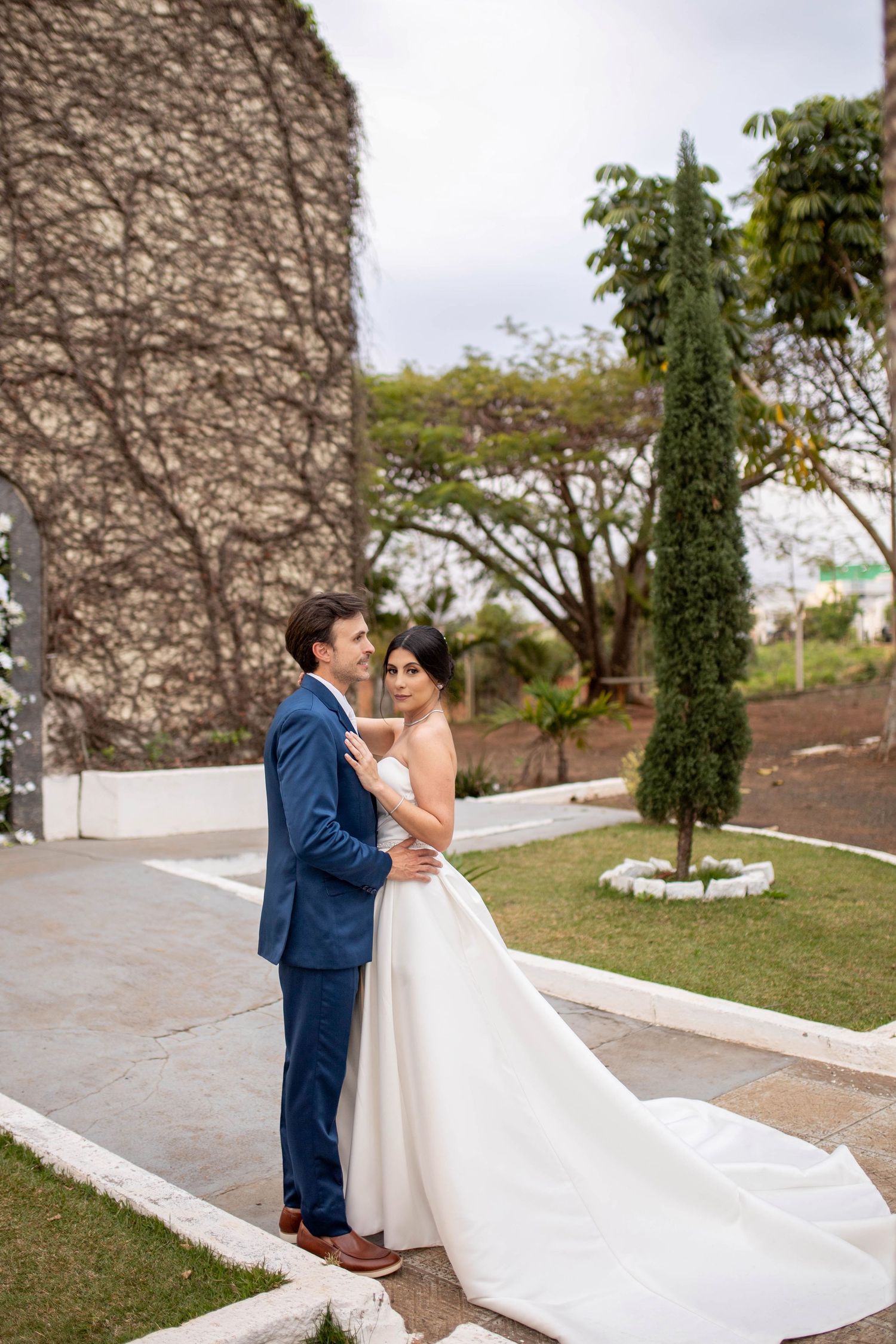 Bride and groom embrace outdoors in a lush garden, wearing a white gown and blue suit on their wedding day.