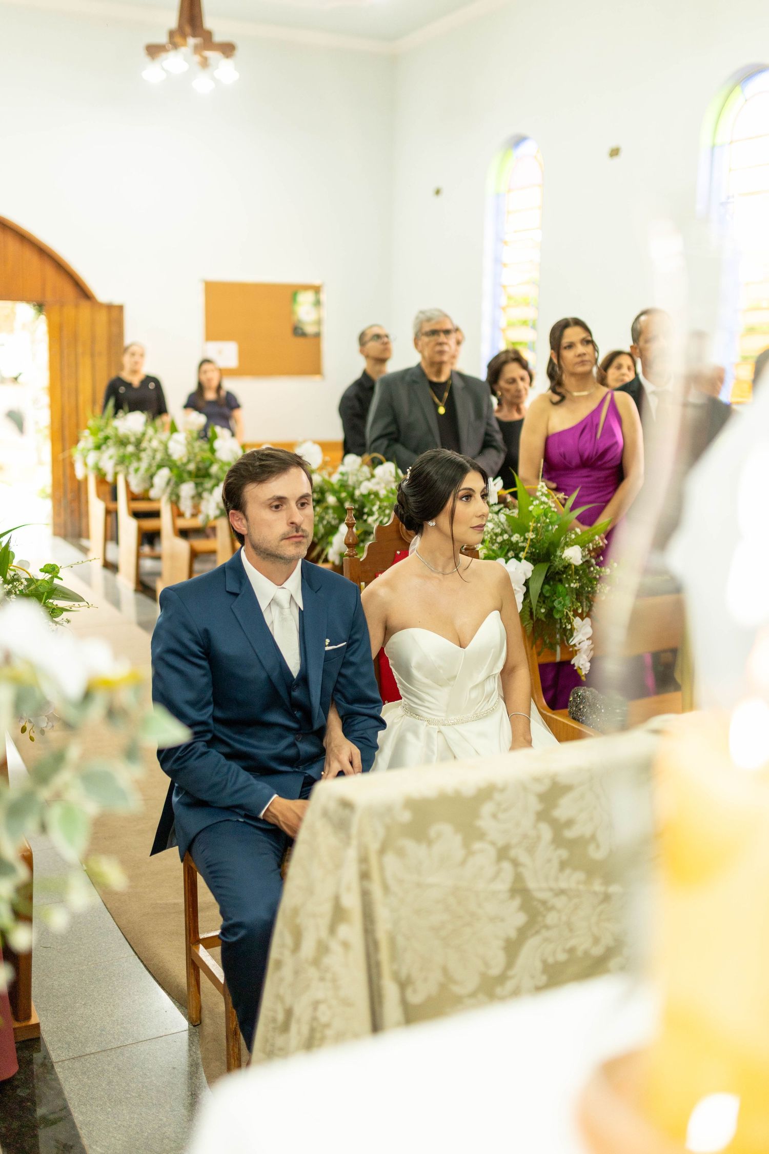 Bride and groom seated at church altar during wedding ceremony with guests in background.