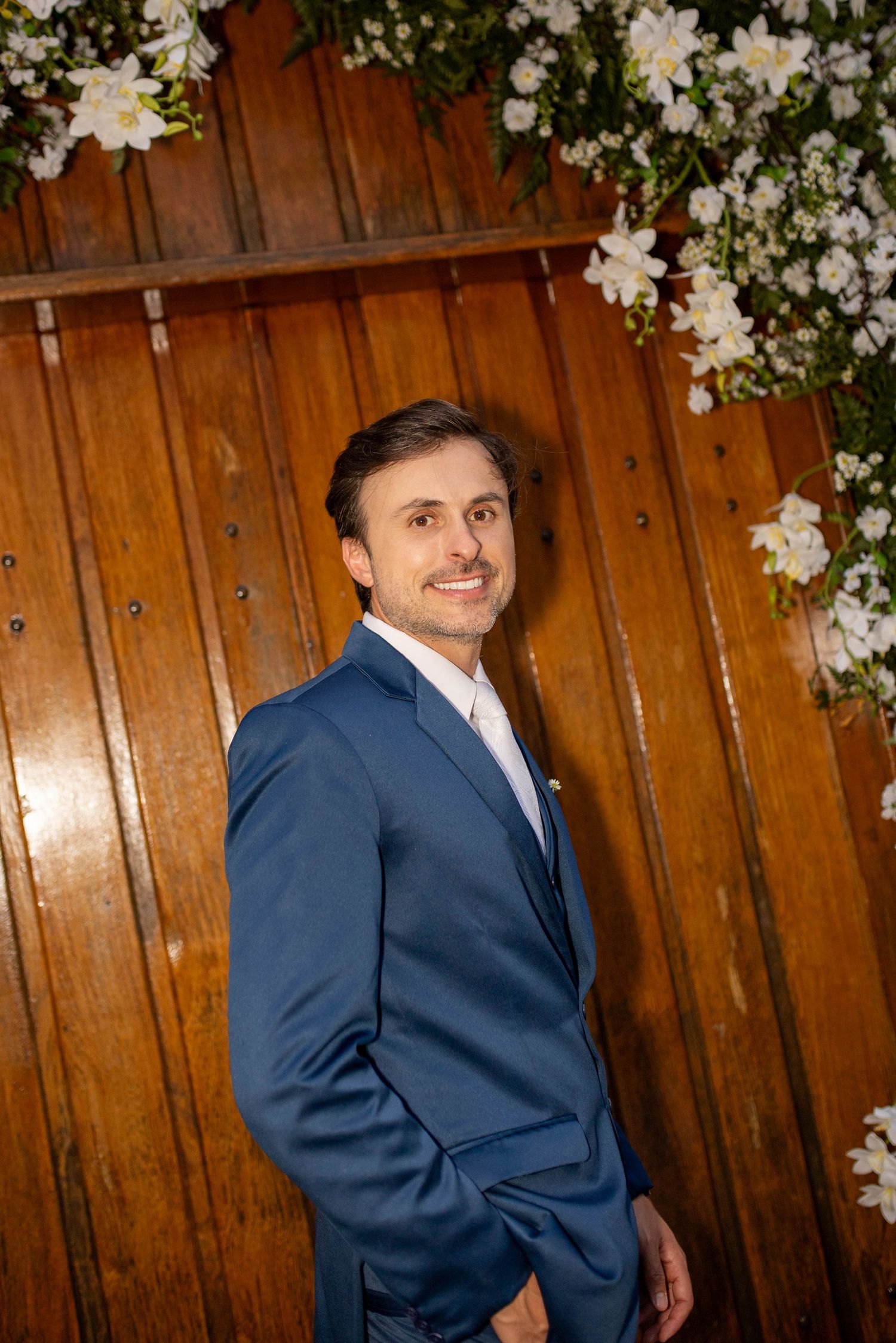 Man in blue suit smiling against wooden wall with white floral decorations at a formal event.