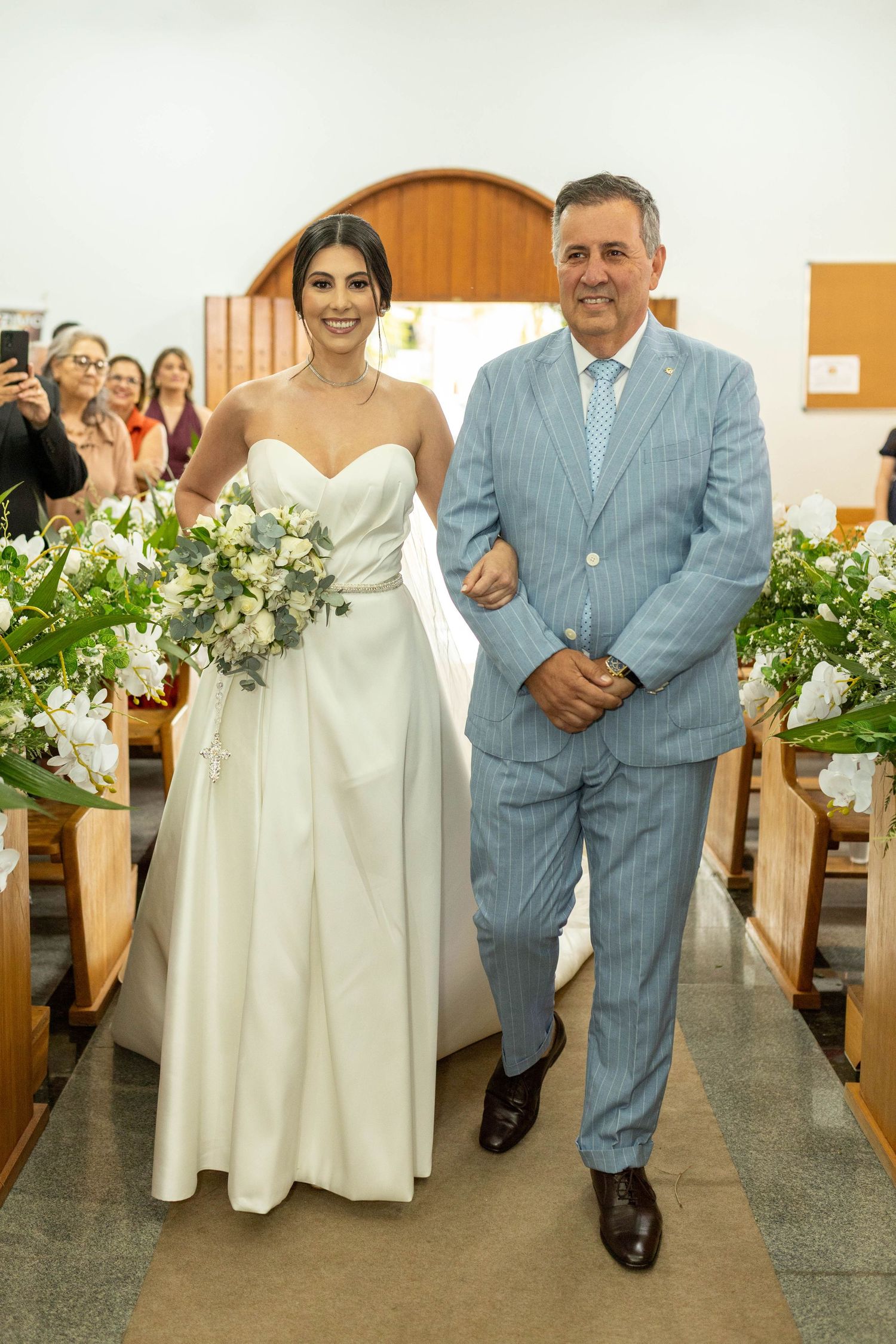 Bride in white strapless gown holding bouquet walks down church aisle with older man in light blue suit.