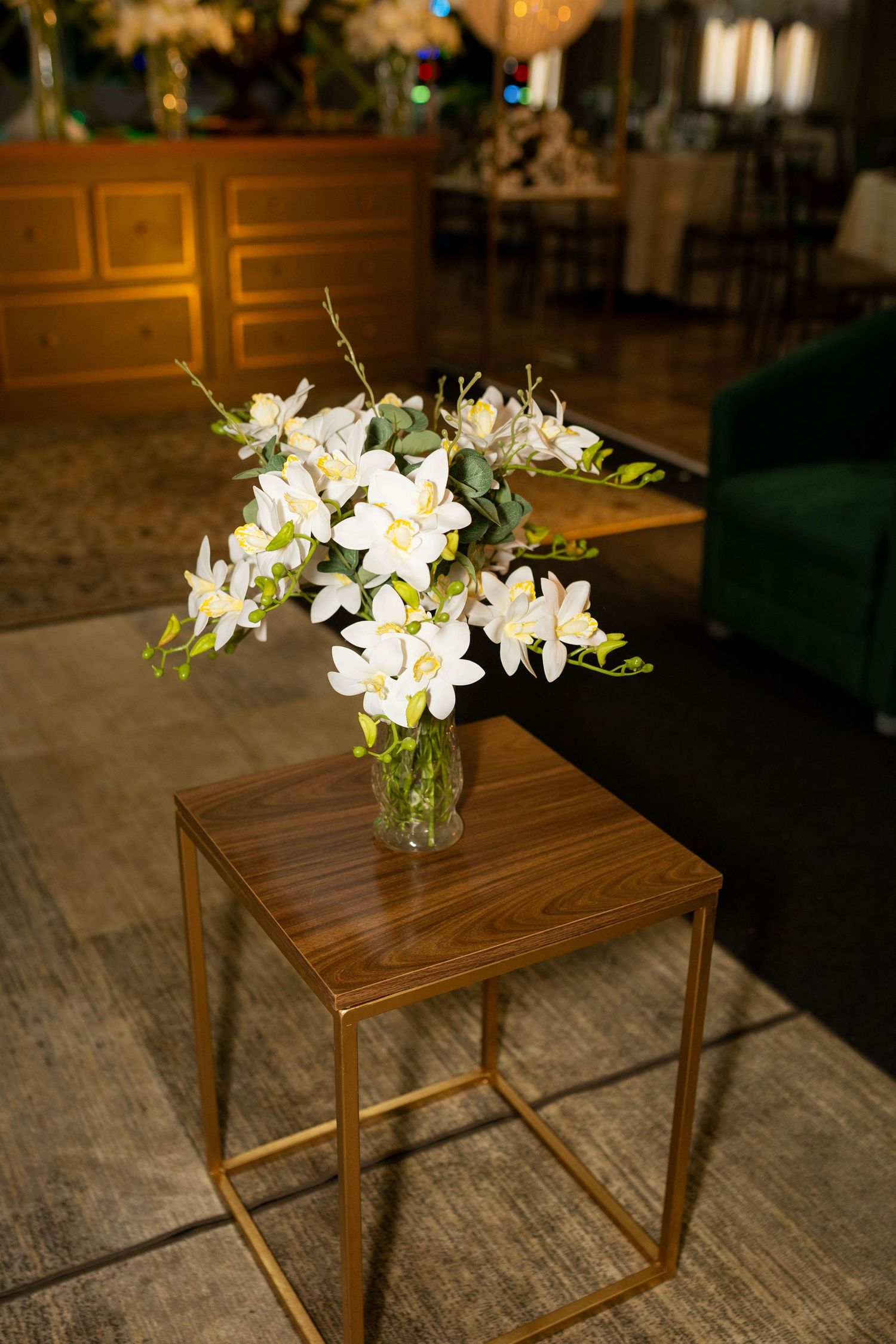 White lily bouquet in glass vase on wooden side table with gold frame in elegant room setting.
