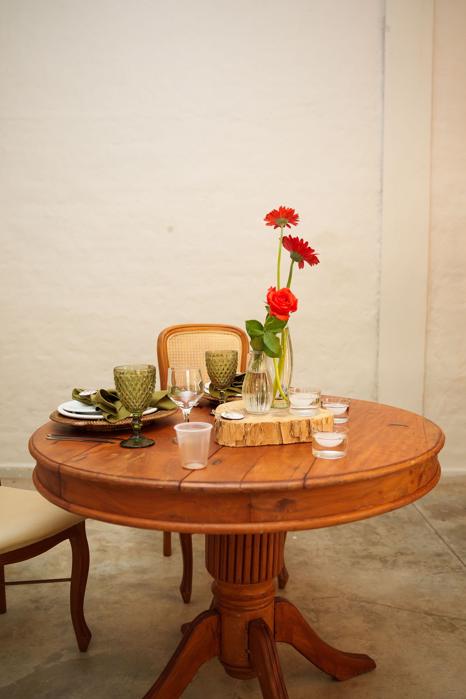 Round wooden pedestal table set with green glasses, plates, and red gerbera daisies in a vase on a wood slice centerpiece.