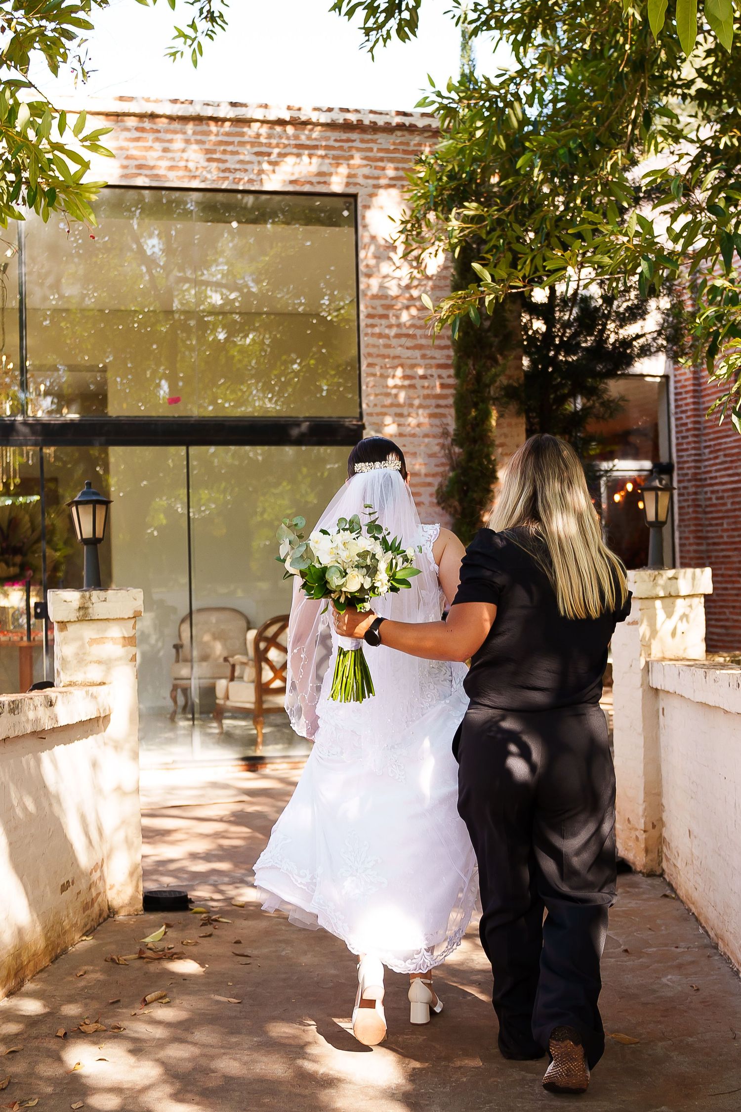 Bride in white gown holding bouquet walks with assistant toward brick building entrance on sunny day.
