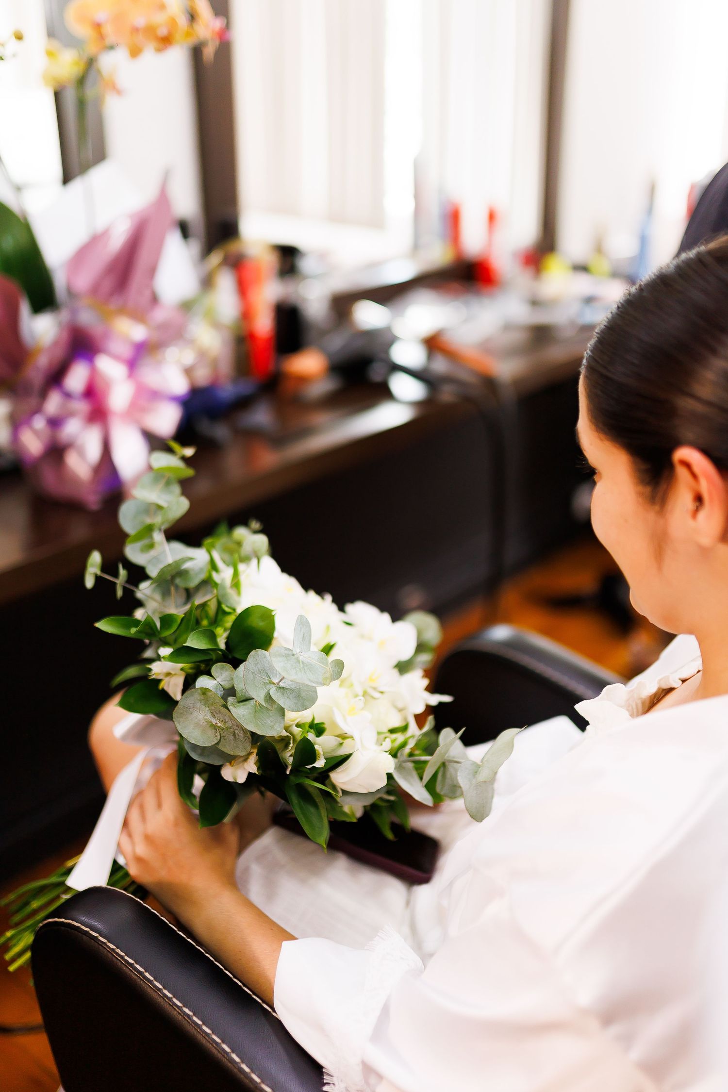 Woman in white shirt arranging a bouquet of white flowers and greenery while seated in a salon chair.