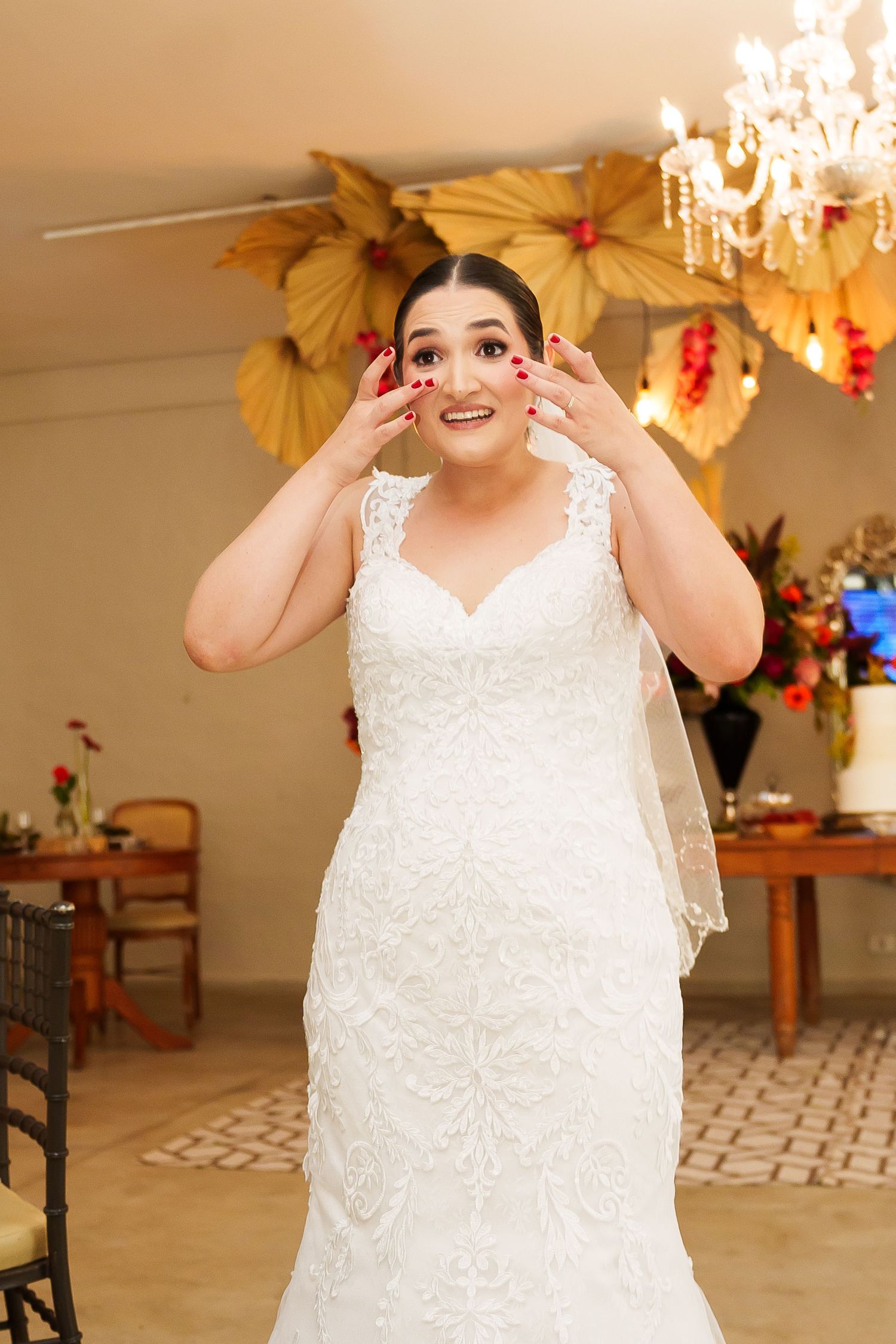 Bride in white lace mermaid gown with veil, hands on cheeks, smiling in decorated wedding venue.