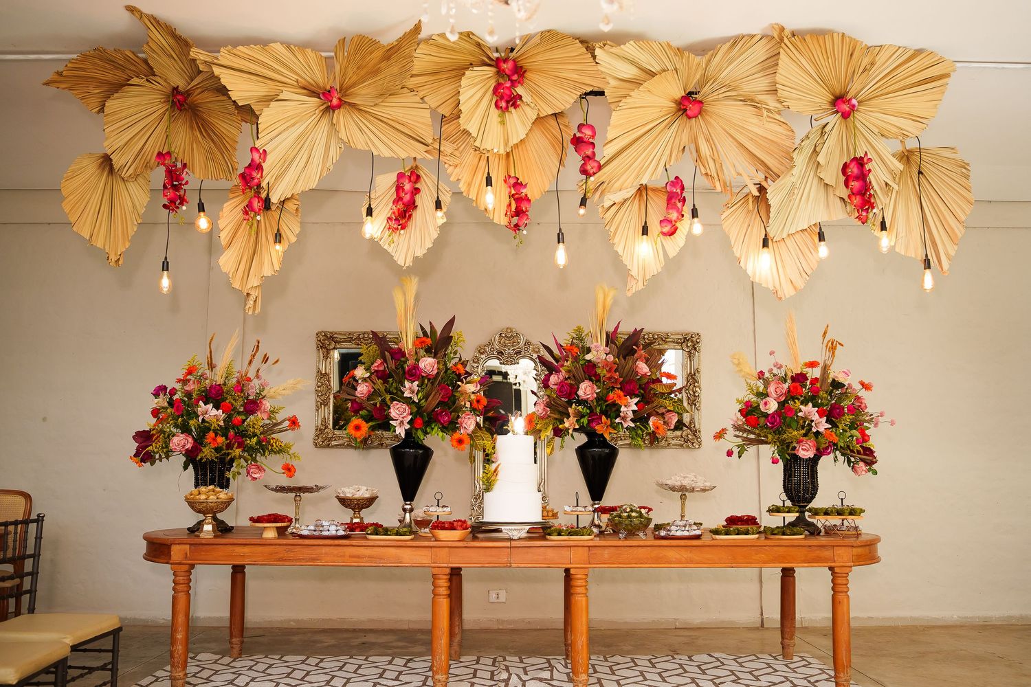 Elegant wedding dessert table with tropical floral arrangements, dried palm leaf canopy, and white cake centerpiece.