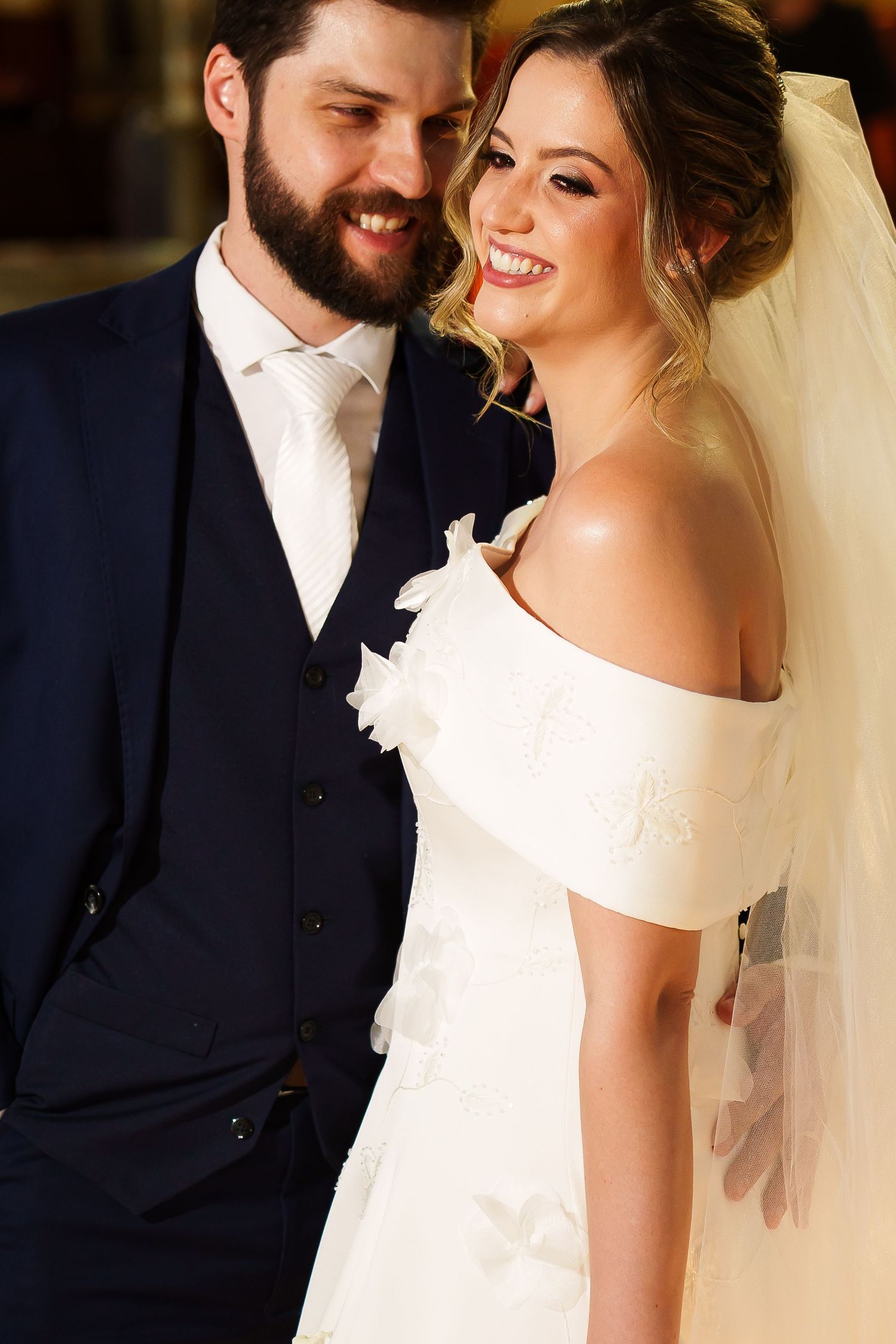 Happy bride in off-shoulder white gown and veil smiles beside bearded groom in navy suit on their wedding day.
