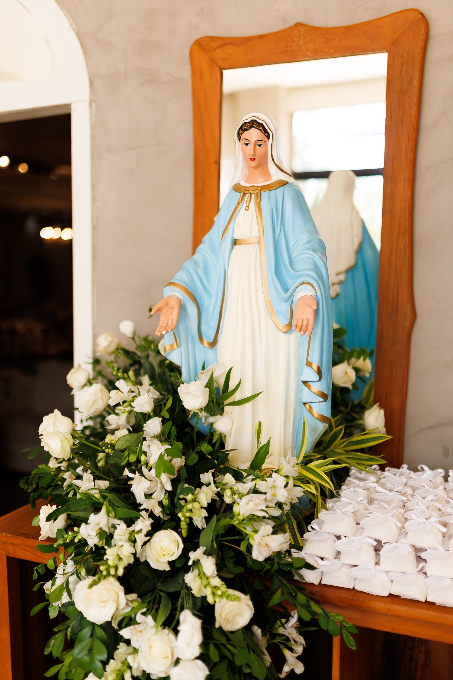 Virgin Mary statue in blue robe surrounded by white roses and flower arrangement in a church setting.