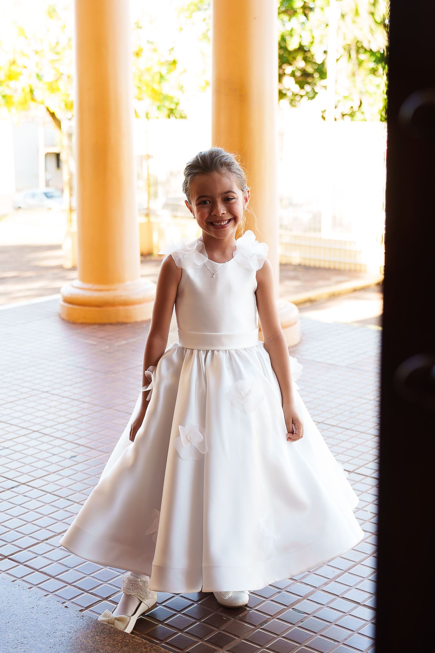 Smiling young girl in white flower girl dress standing between yellow columns at outdoor venue.