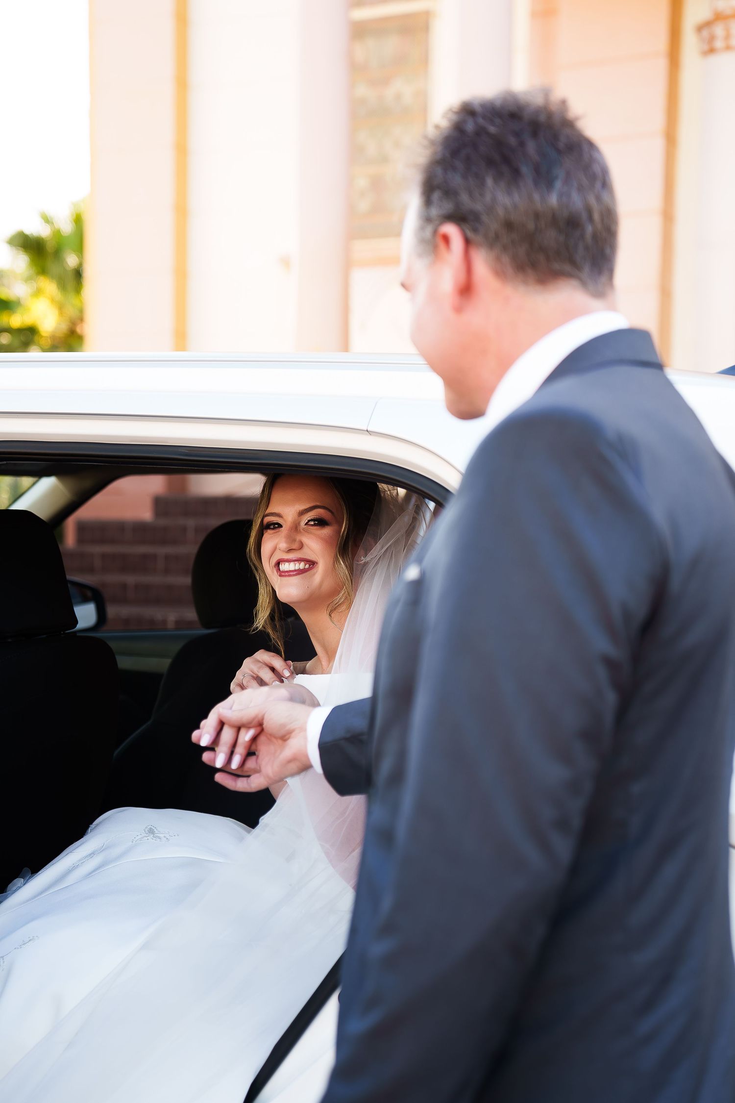 Smiling bride in white dress holds hands with groom in gray suit while sitting in a white wedding car.