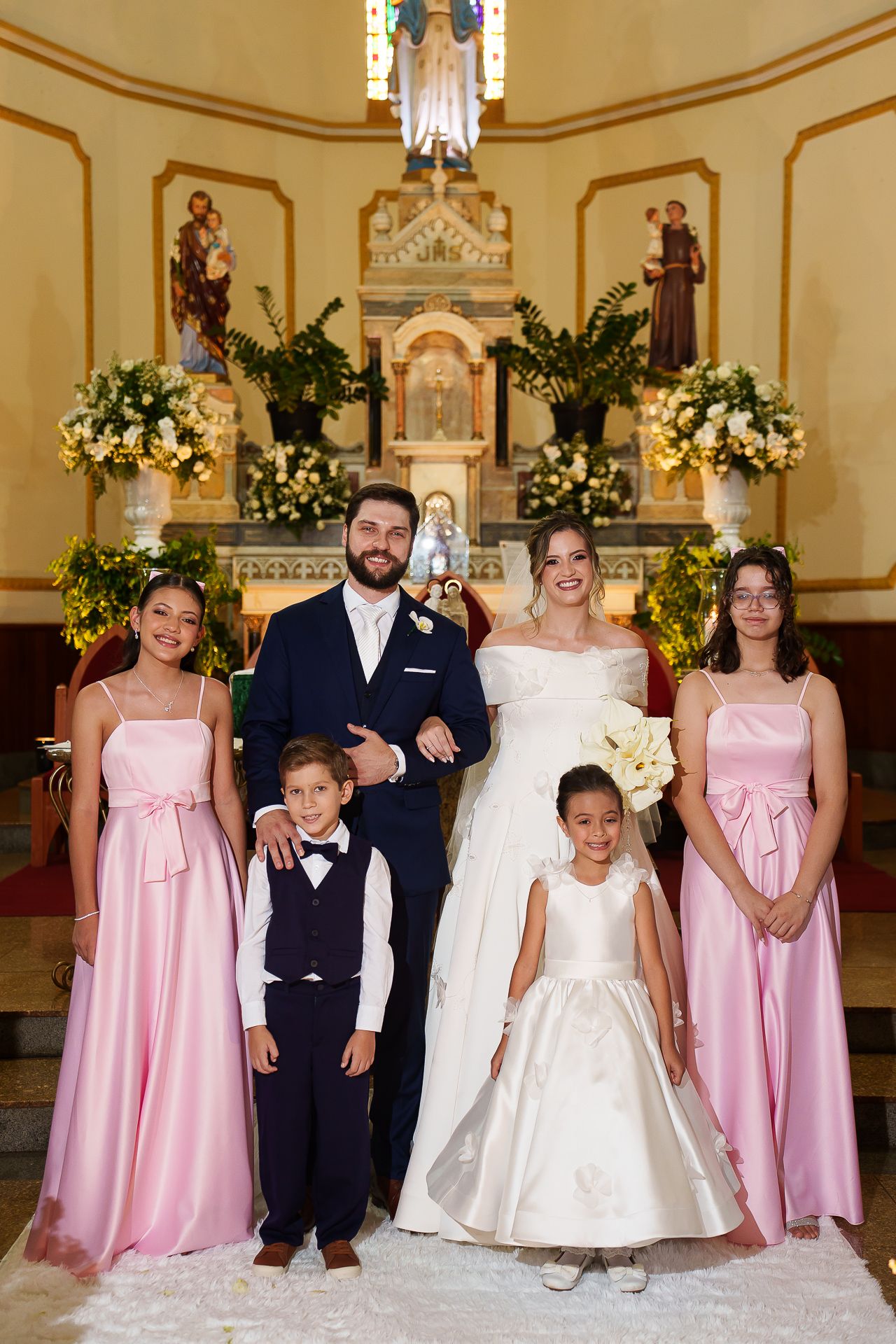 Bride and groom pose with bridesmaids and children at church altar decorated with white flowers.