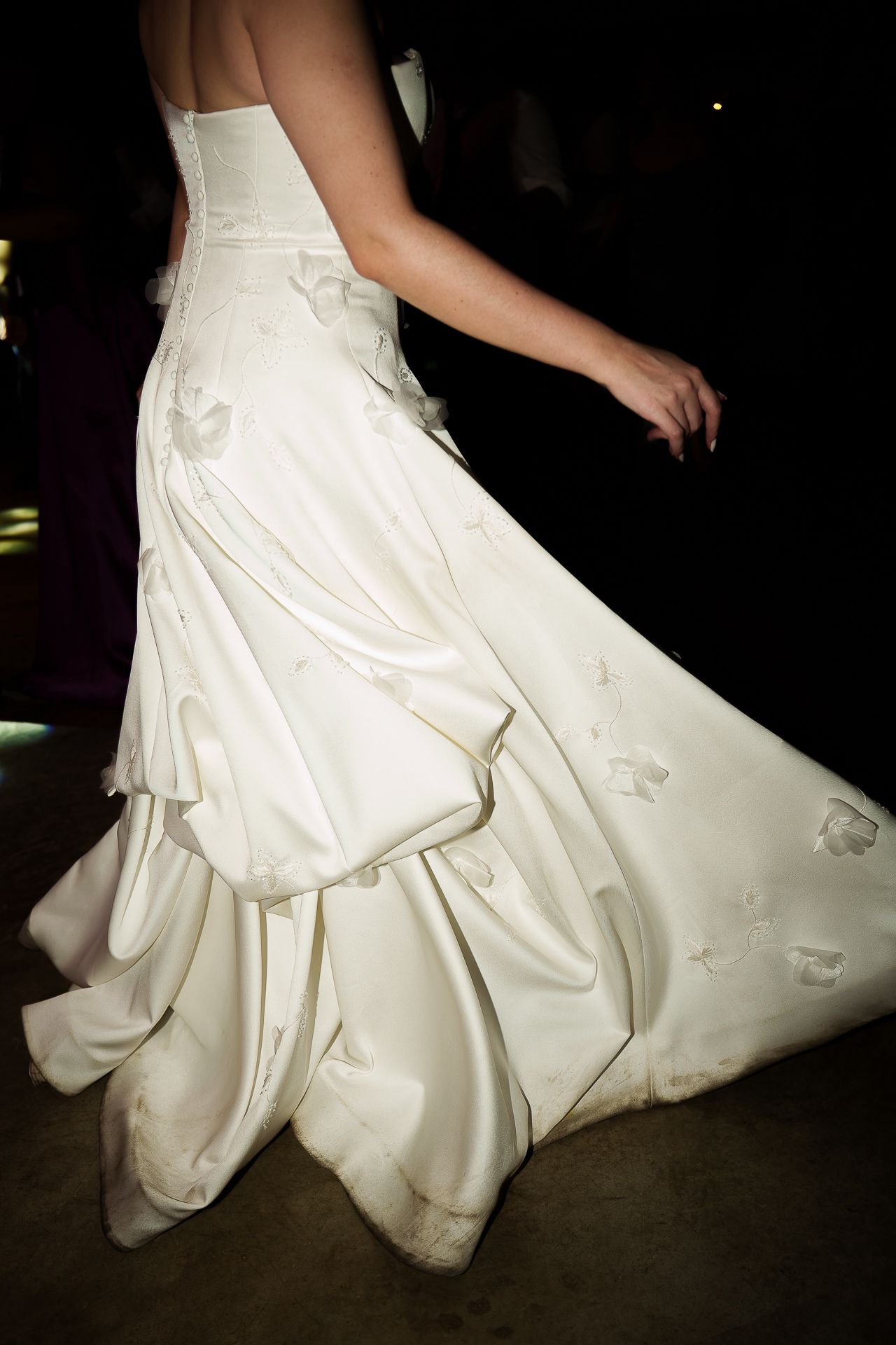 Elegant bride in a flowing white satin wedding gown with floral appliques, captured mid-movement on a dark dance floor.