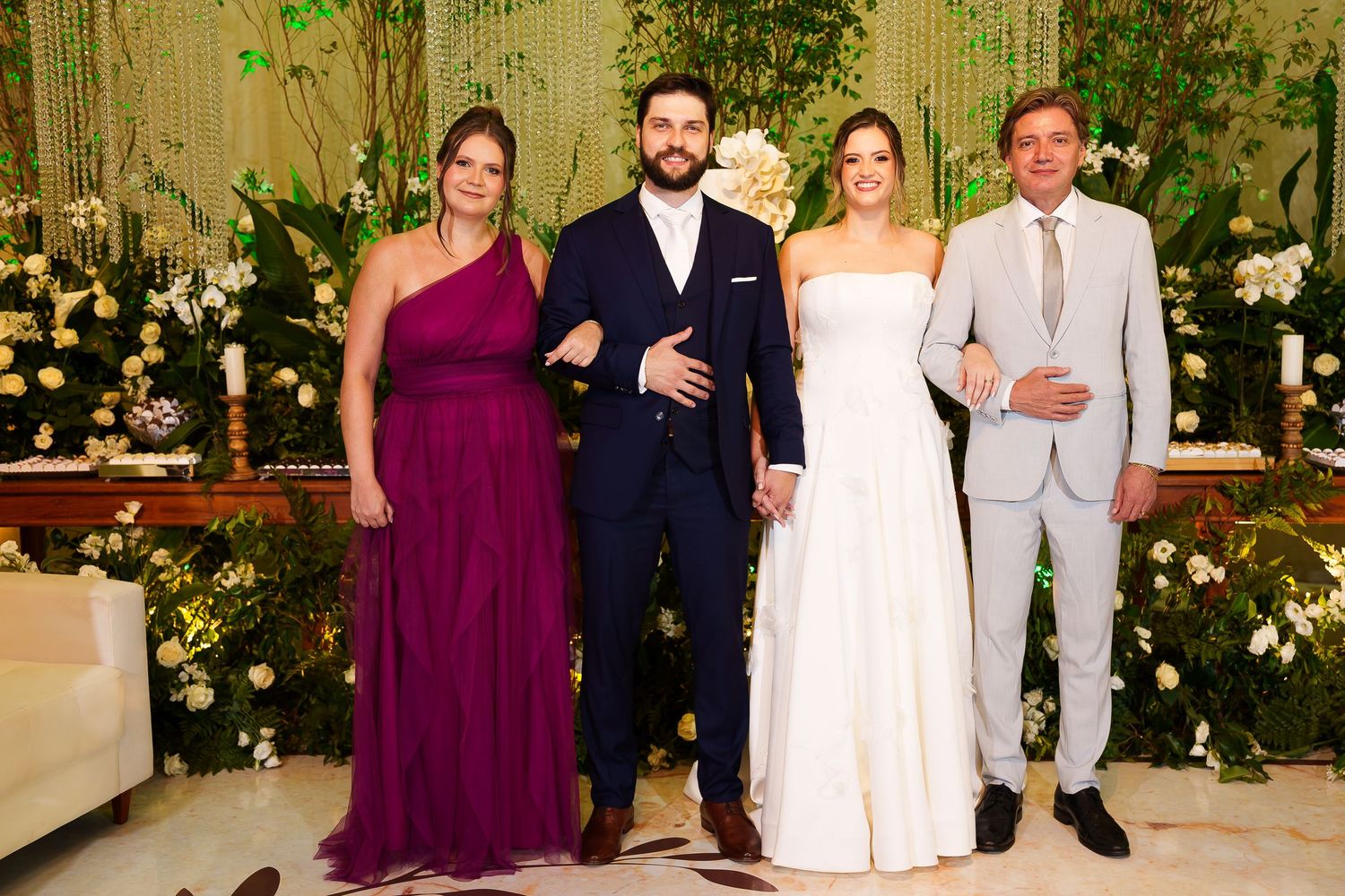 Elegant wedding party of four posing in front of floral backdrop, bride in white gown, groom in navy suit.