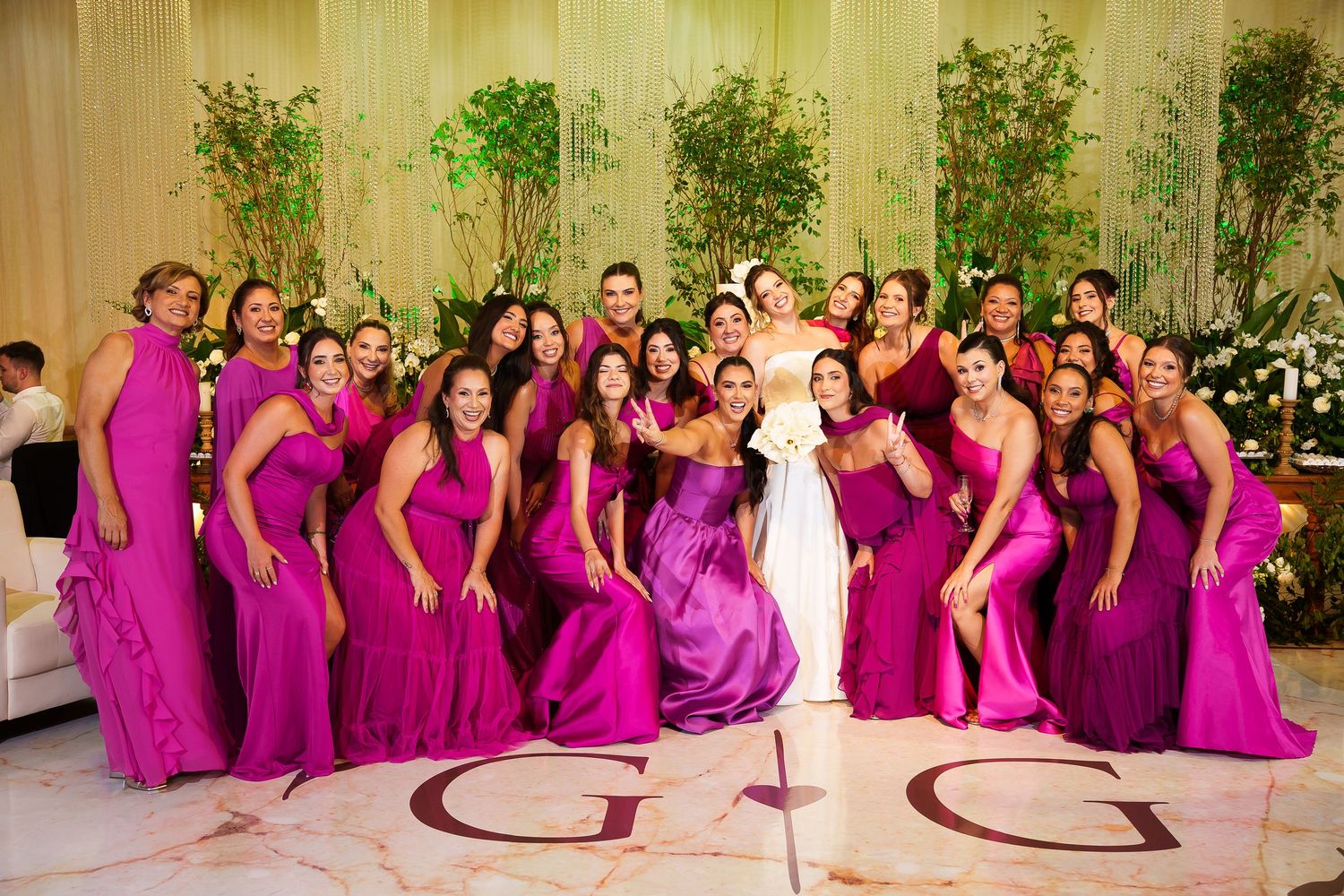 Bride surrounded by bridesmaids in fuchsia gowns posing on dance floor with monogram at elegant wedding reception.