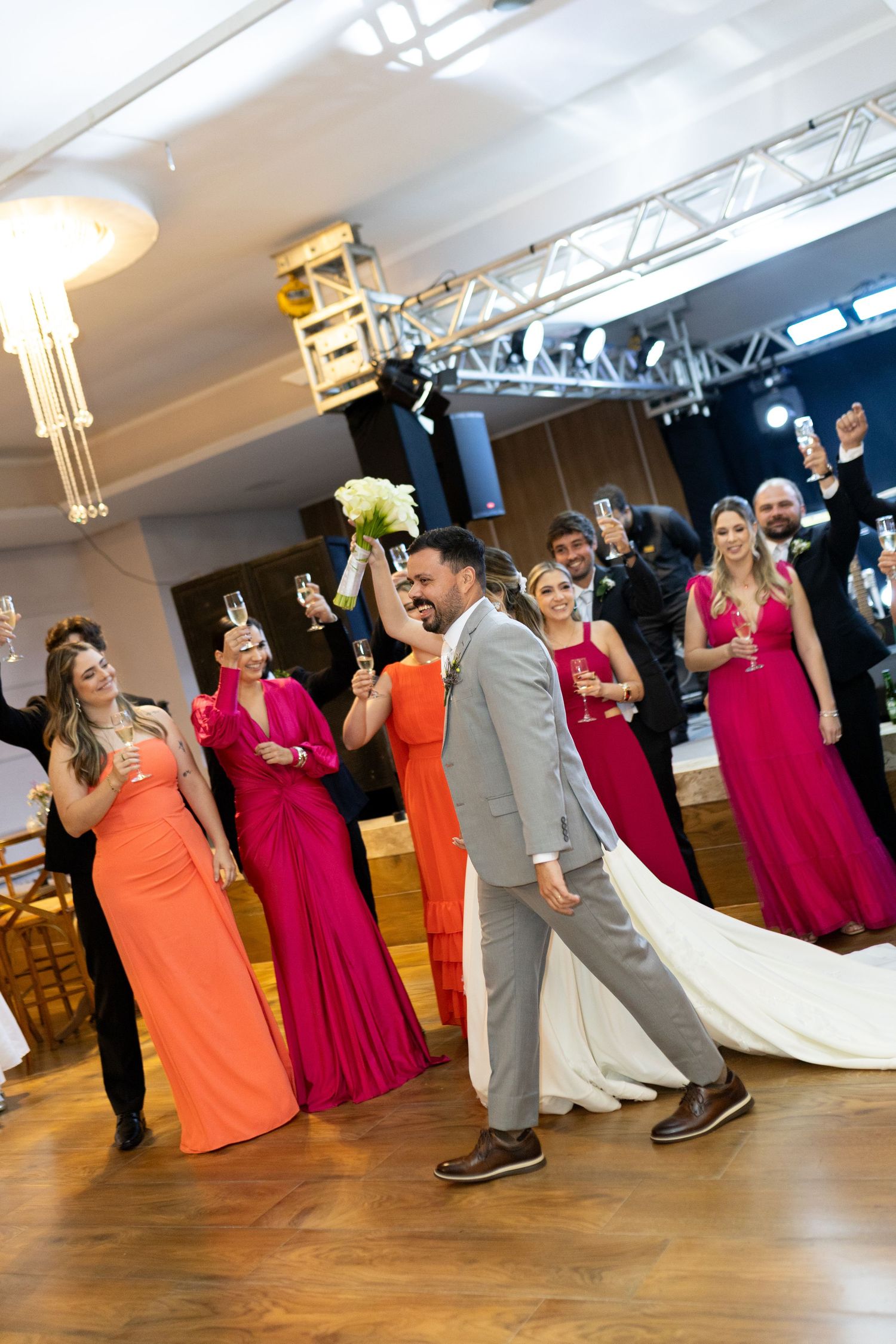 Bride and groom walk together as bridesmaids in pink and orange gowns raise champagne glasses in celebration.