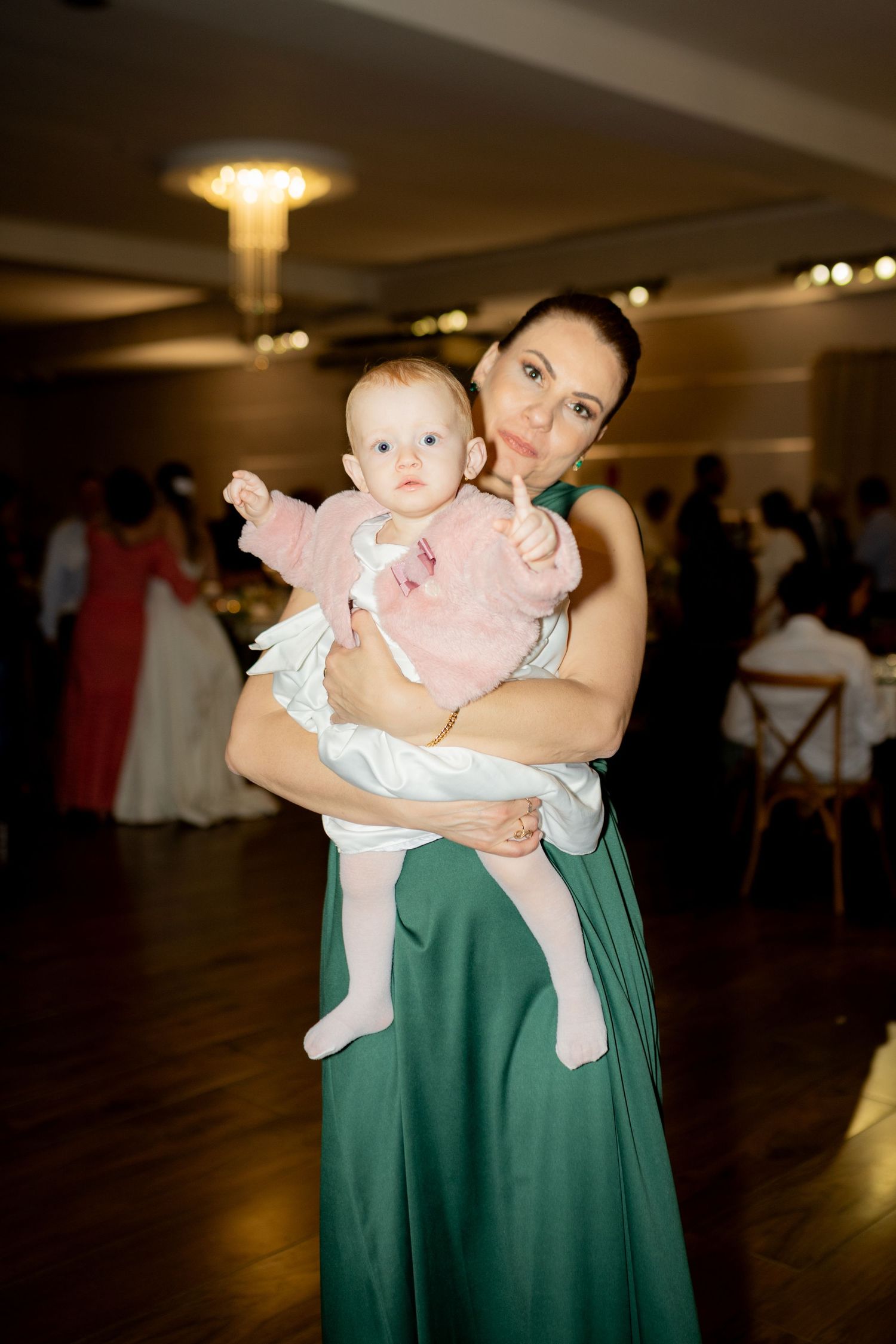 Woman in green gown holds baby in pink outfit at elegant wedding reception venue.