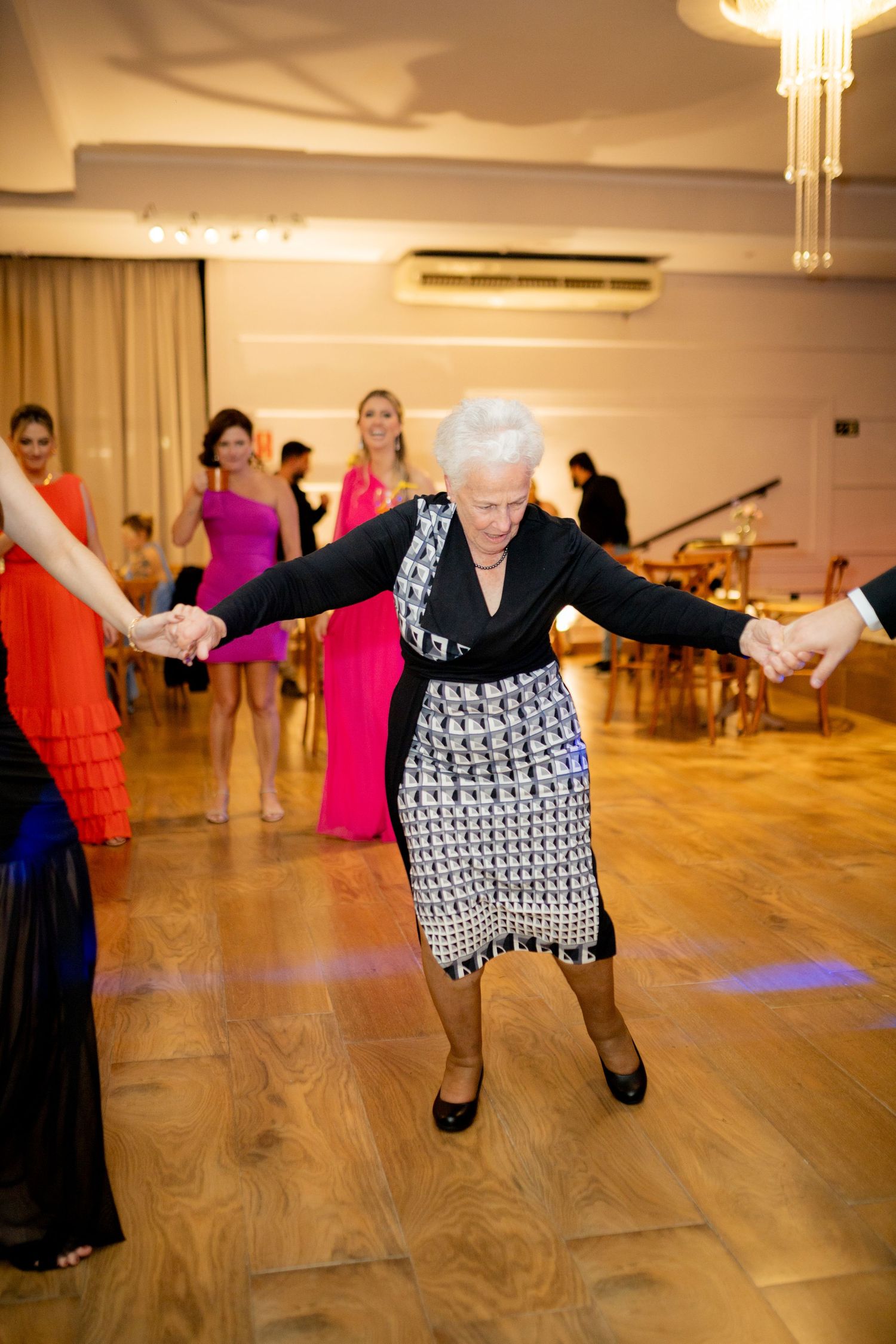 Elderly woman dancing joyfully at a formal event, holding hands with others on a ballroom dance floor.