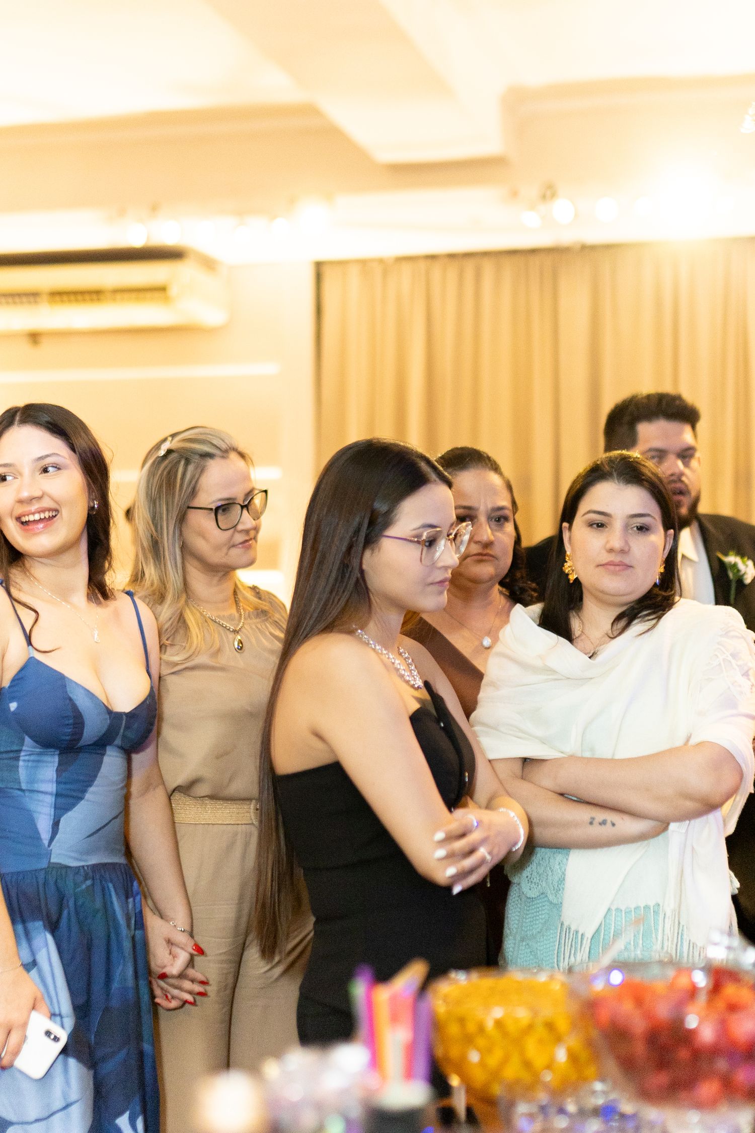 Group of women standing together at an indoor event, smiling and watching a presentation in a warmly lit room.