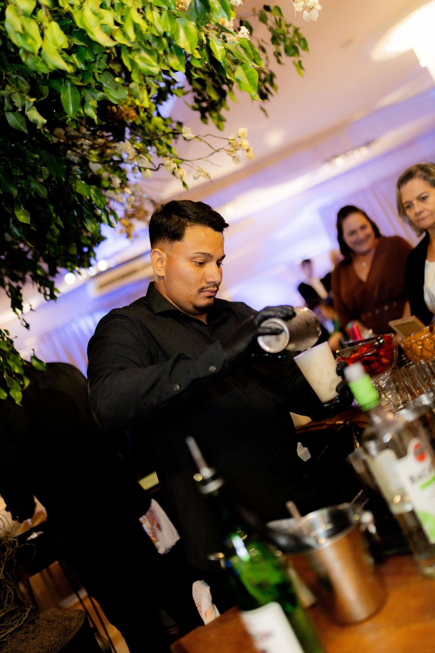 Bartender in black uniform mixing cocktails at an elegant event with green foliage decor and guests in background.
