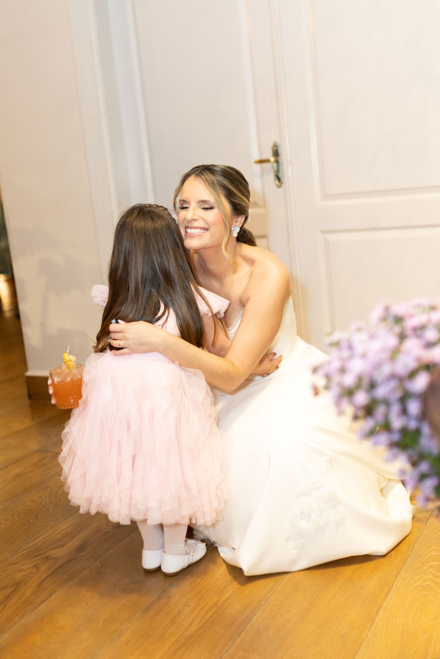 Bride in white gown kneels to hug a little girl in a pink tutu dress at a wedding reception.