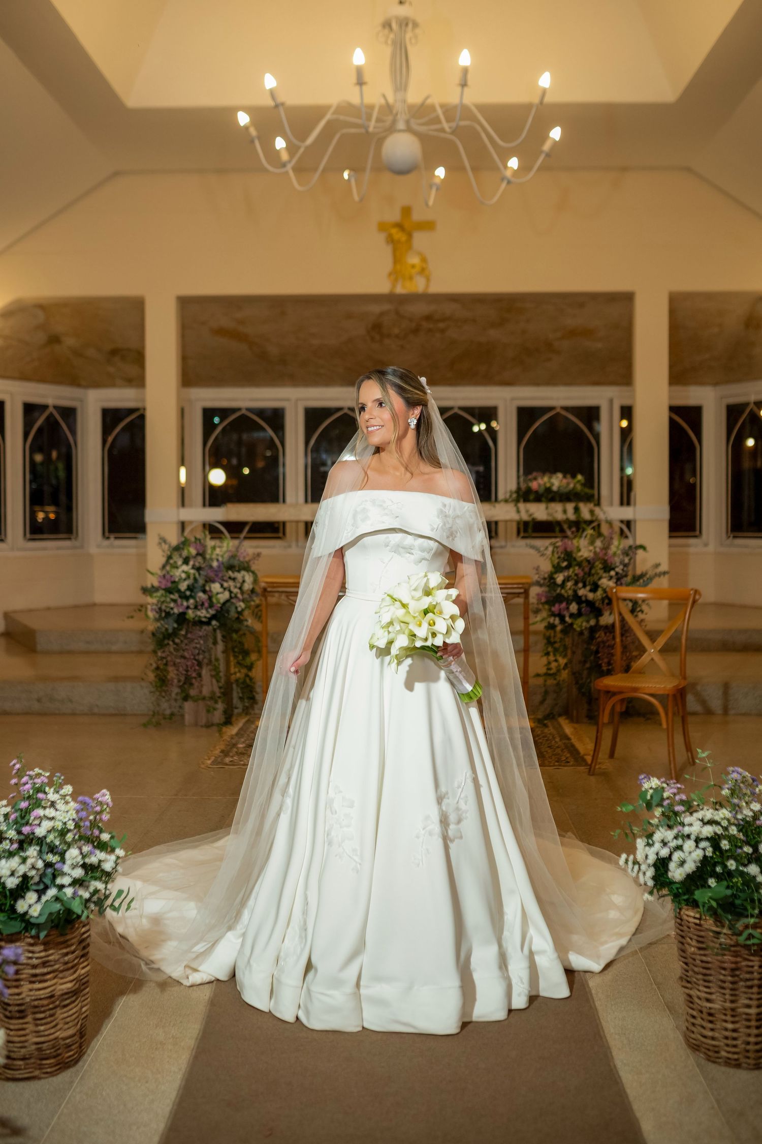 Bride in off-shoulder white gown with veil holds bouquet in elegant chapel with chandelier and floral arrangements.