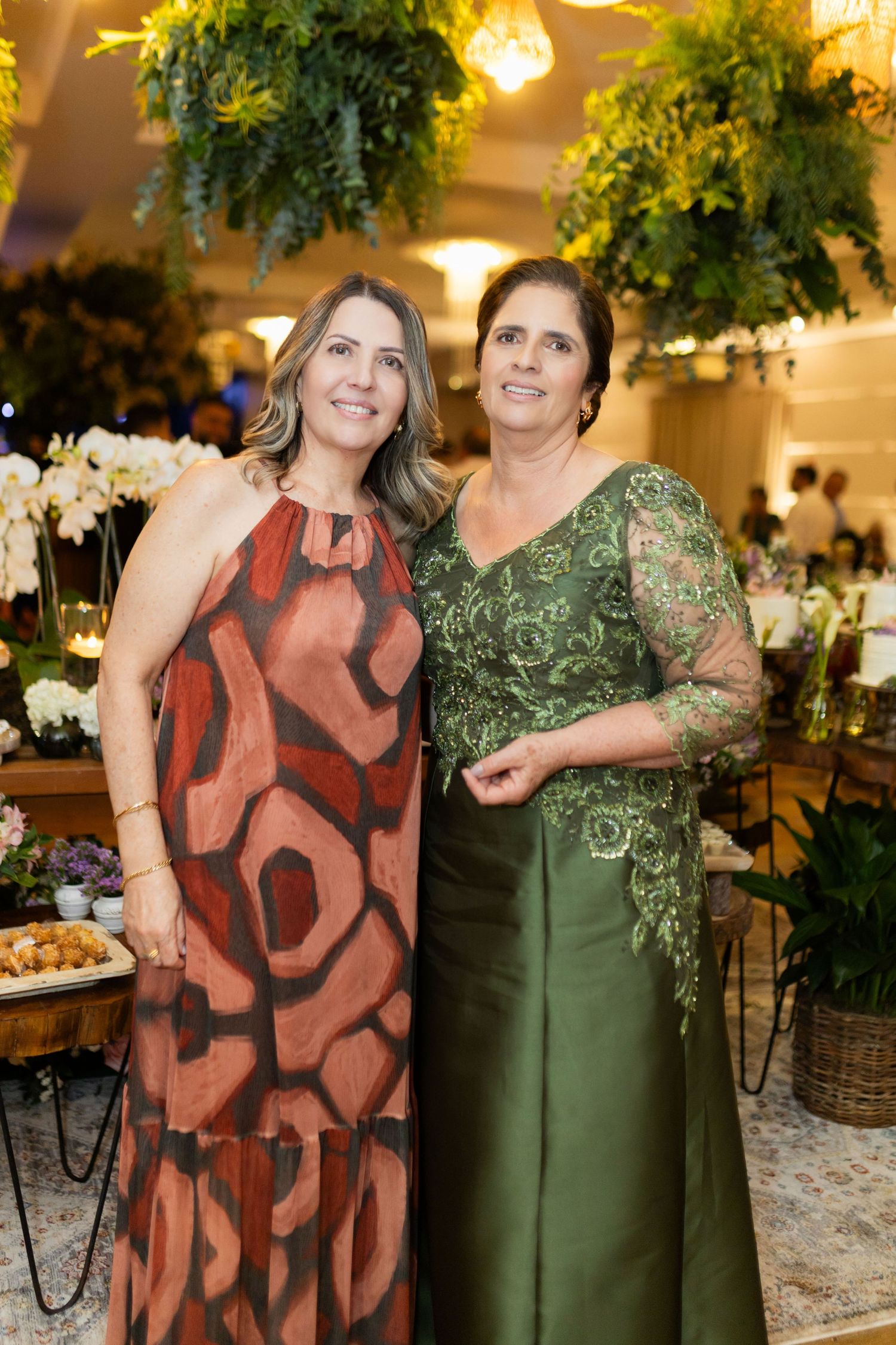 Two women smile together at an elegant event, one in a red patterned dress, the other in a green lace gown, surrounded by floral decor.