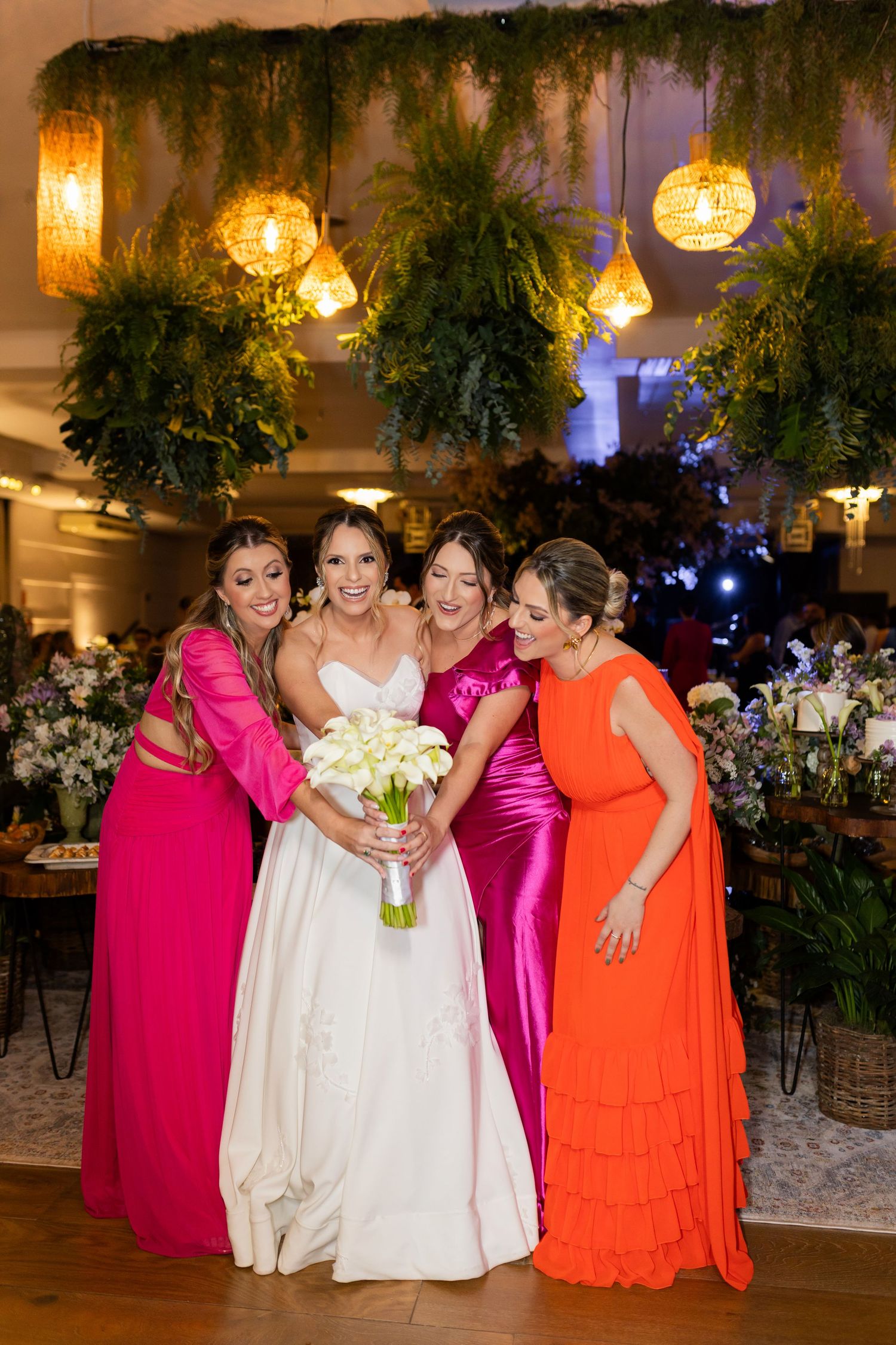 Bride in white gown holds calla lily bouquet with three bridesmaids in pink and orange dresses at elegant wedding reception.