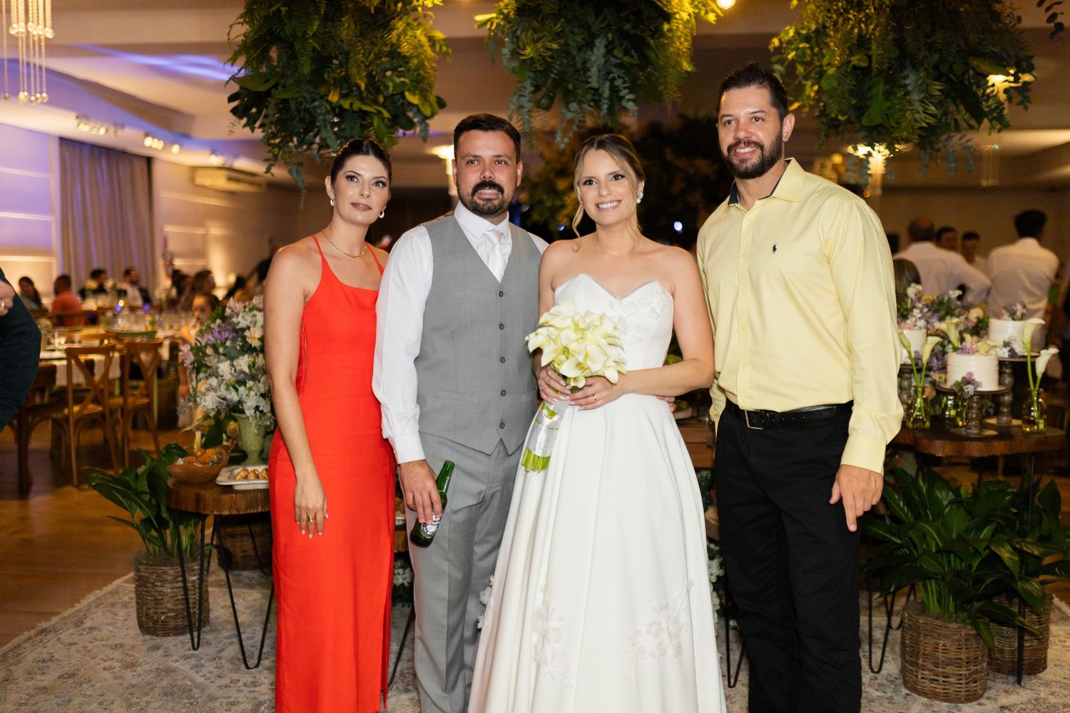 Bride in white gown and groom in gray vest pose with two guests at elegant wedding reception venue.