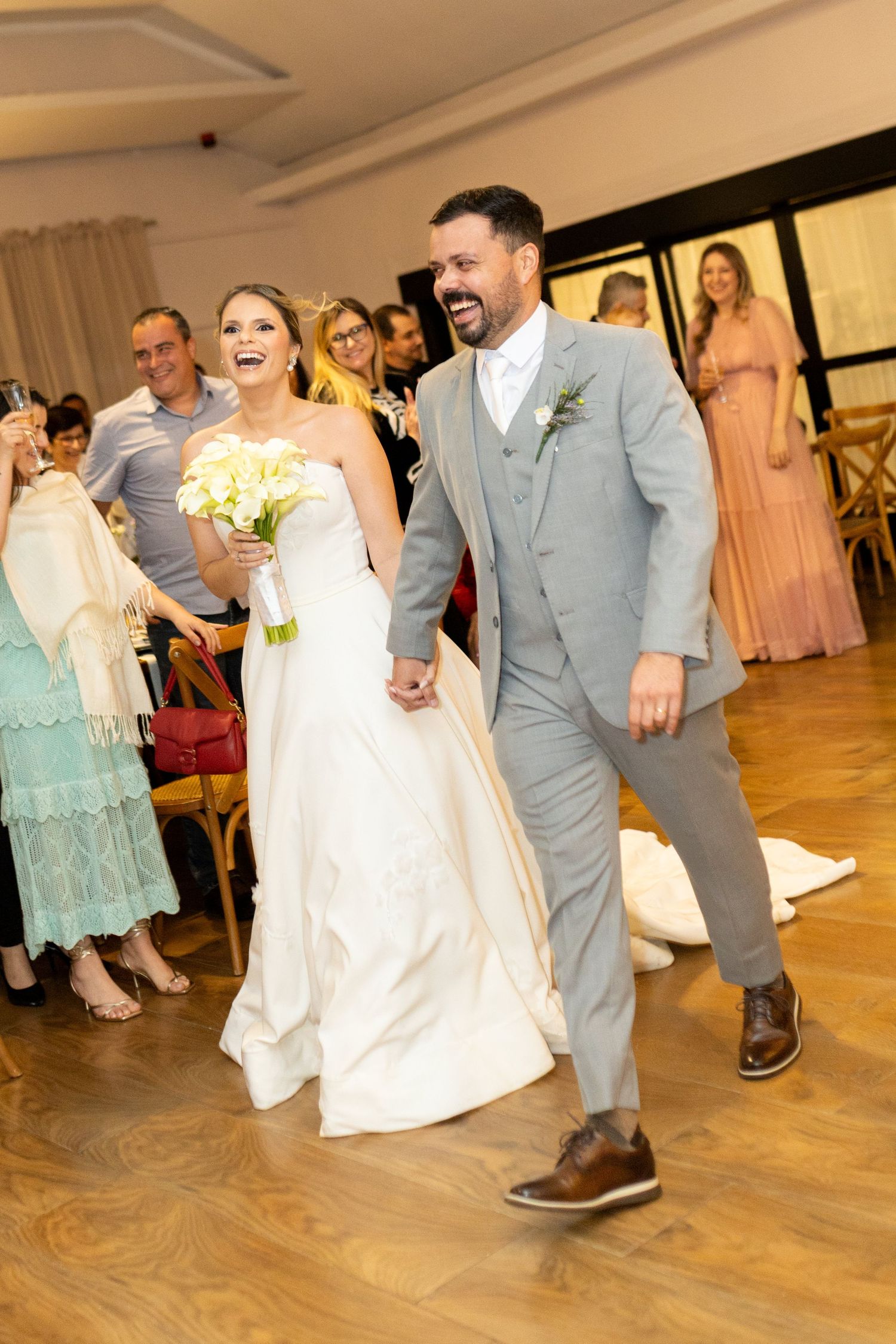 Joyful newlywed couple walks hand-in-hand at wedding reception, bride in white gown holding bouquet, groom in gray suit.