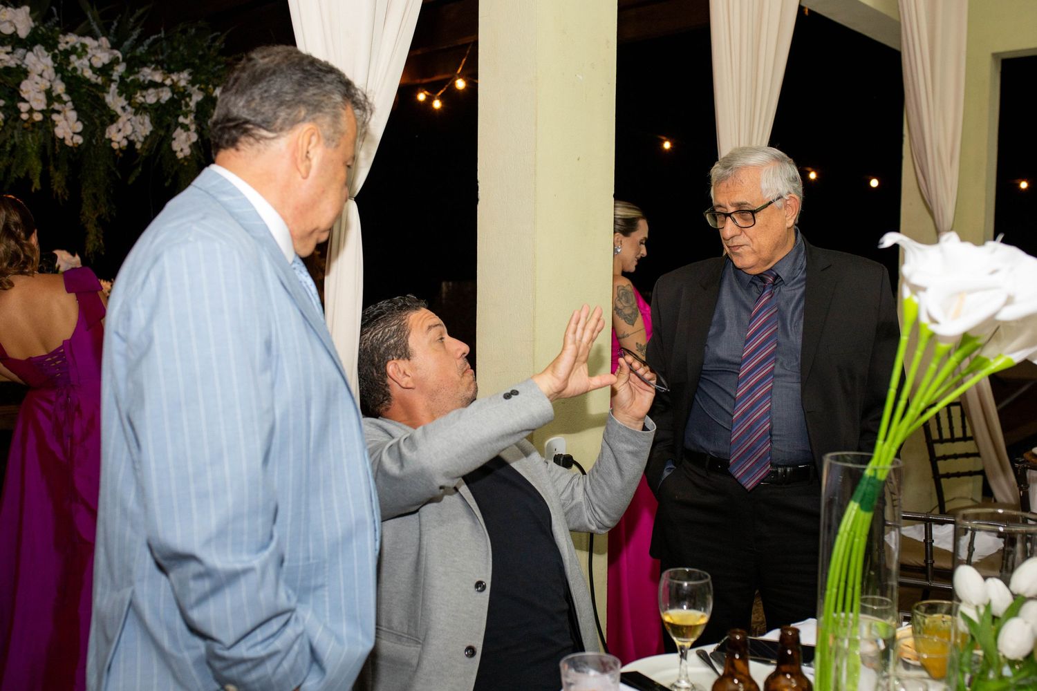 Three men in formal attire interact at an elegant evening event, with one man giving a high five.