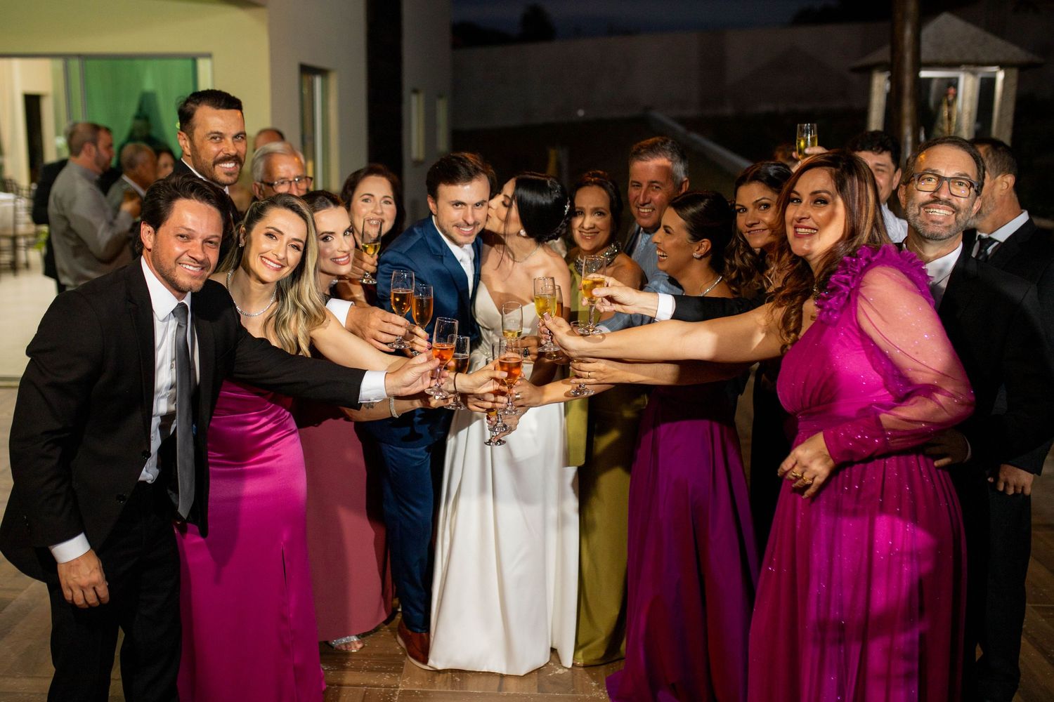 Wedding guests in purple and pink dresses toast champagne with bride and groom at elegant outdoor reception.