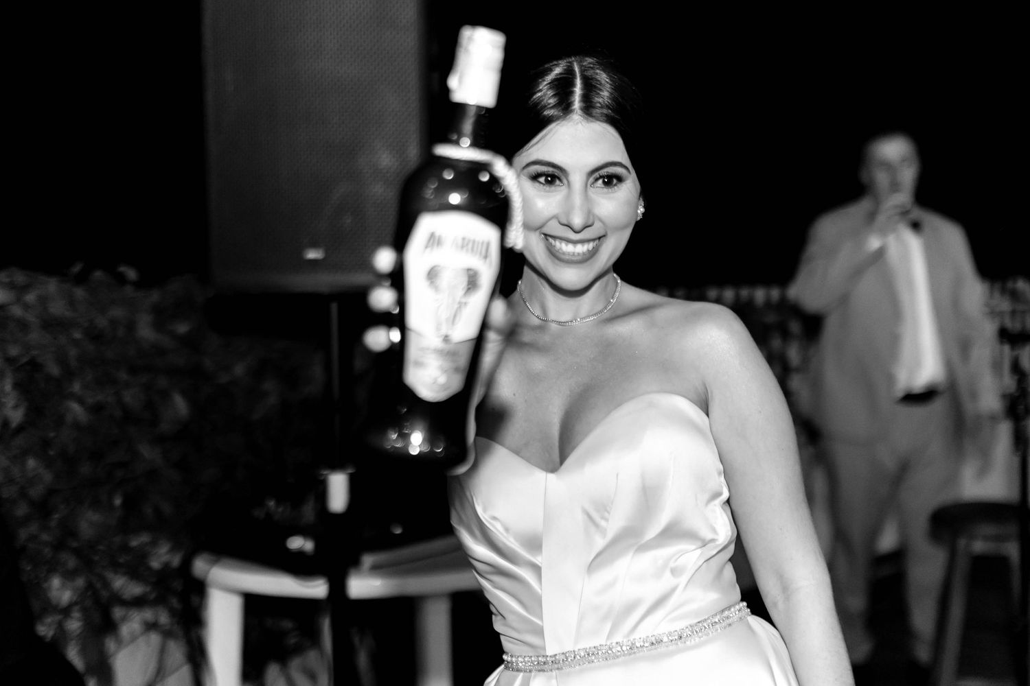 Bride in strapless gown smiling and holding up a bottle of liquor at a wedding reception in black and white.